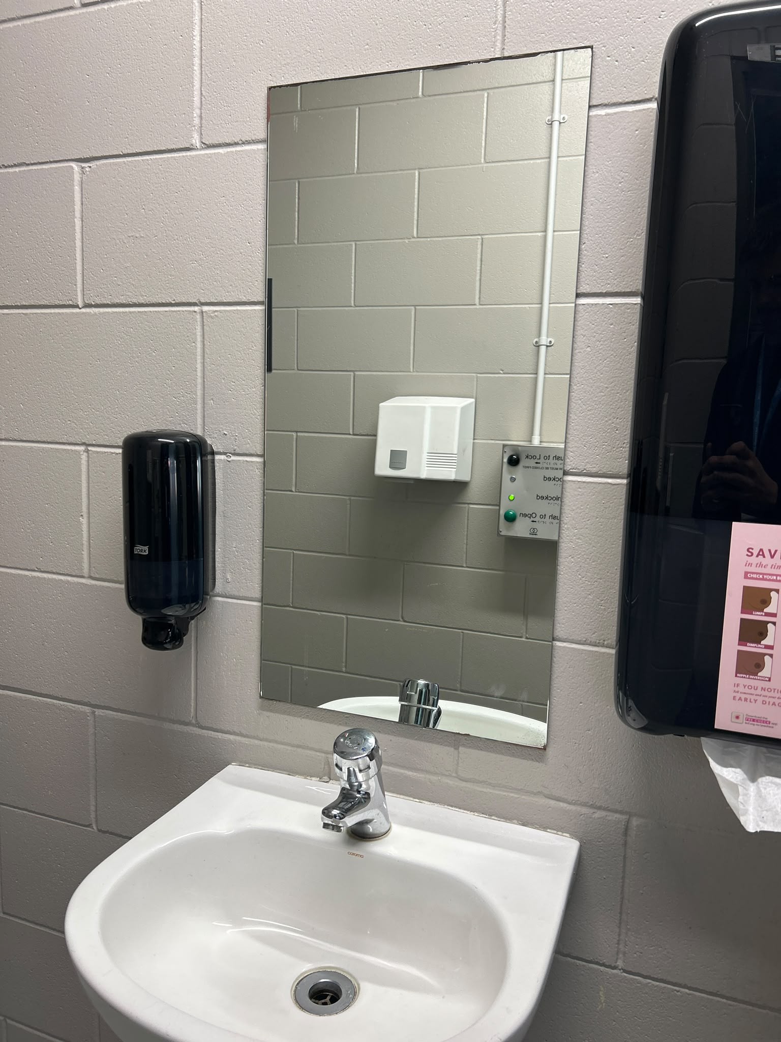 White washbasin with single lever tap on a grey block wall. A mirror reflects door locking controls and a hand dryer. Black soap and paper towel dispensers are mounted on the wall.