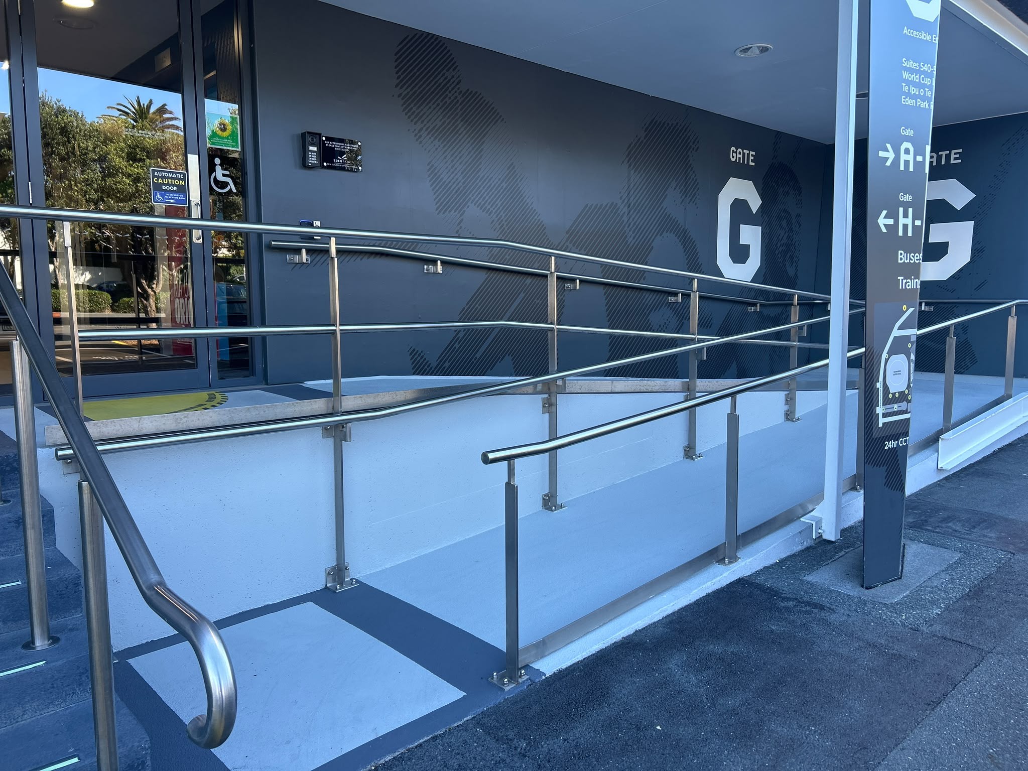 Light grey concrete ramp with dual stainless steel handrails leading to an automatic glass door. Large white 'Gate G' text on dark grey wall and vertical signage for accessible transport options.