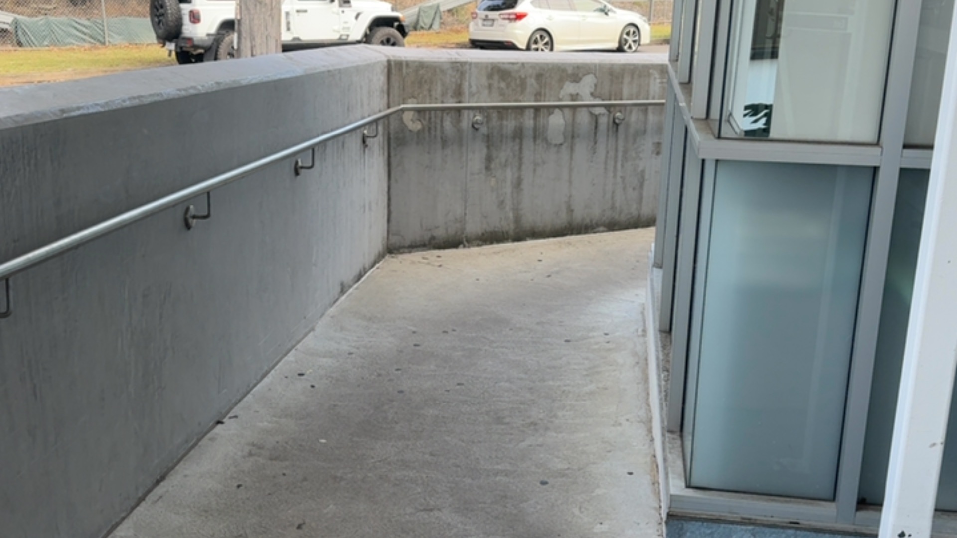 Sloping concrete ramp with metal handrails along a grey concrete wall, leading towards a glass-panelled entrance. The ramp has a smooth surface and gradual incline.