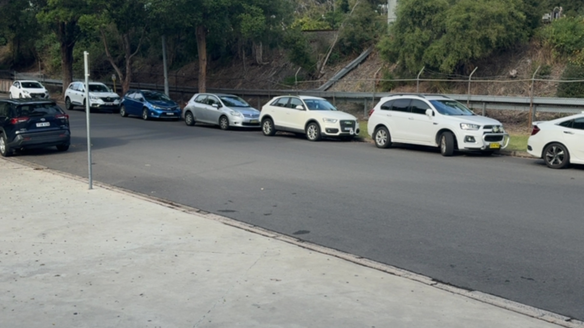 Asphalt road with a level concrete footpath. Several cars parallel parked along the far side under trees. A tall metal signpost stands on the near side.