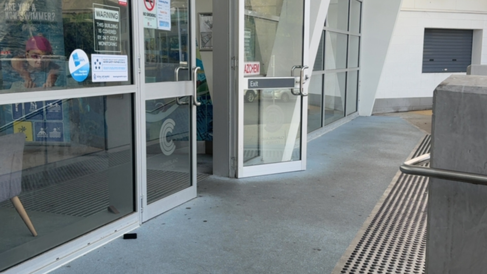 Level entrance with glass double doors and metal handles. Blue non-slip flooring leads to the doorway. A ramp to the right features a metal handrail and tactile ground surface indicators.