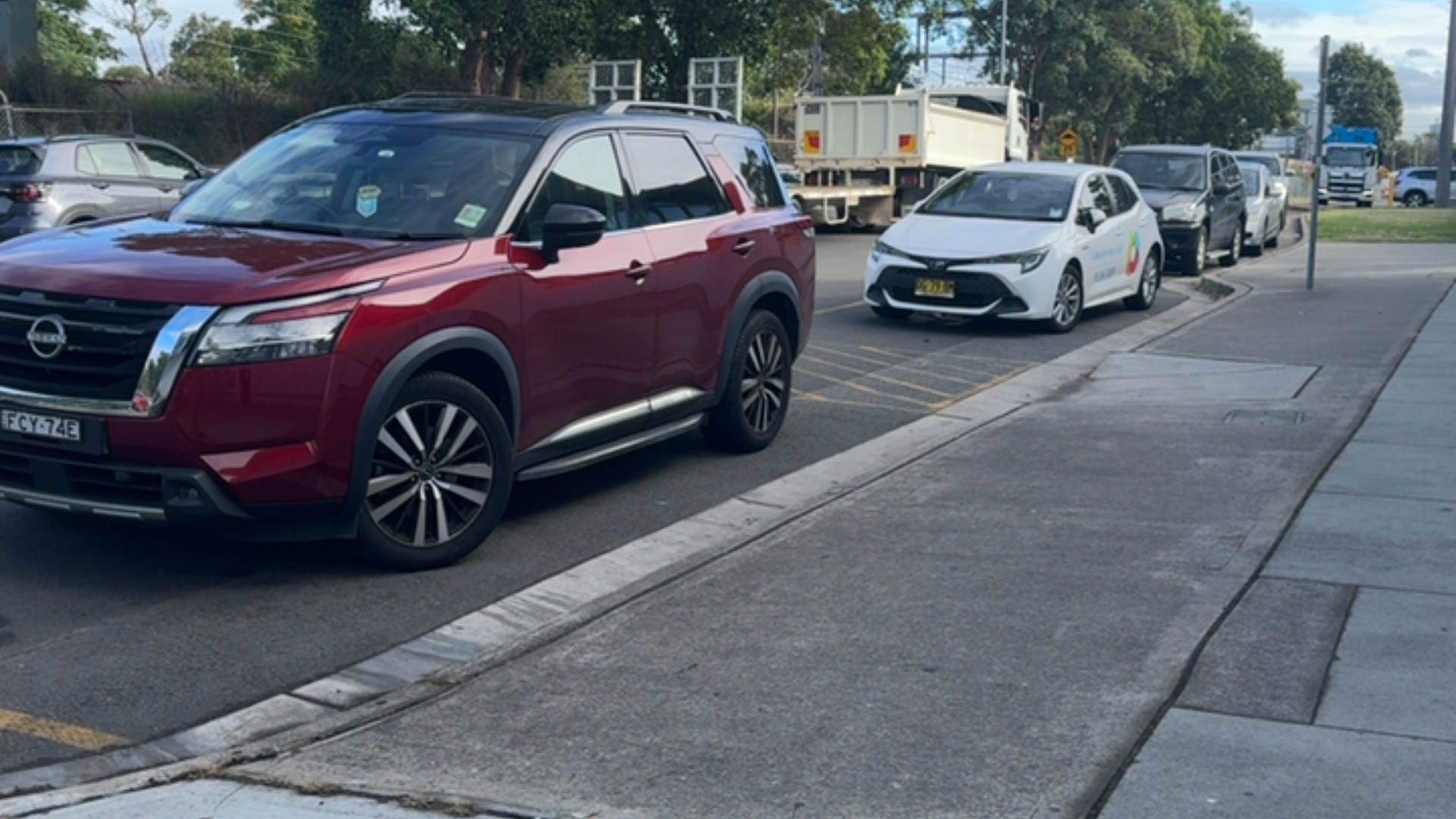 Wide, level concrete footpath with a grey textured surface and a raised concrete kerb. The path is unobstructed and runs parallel to a bitumen road with several parked vehicles.