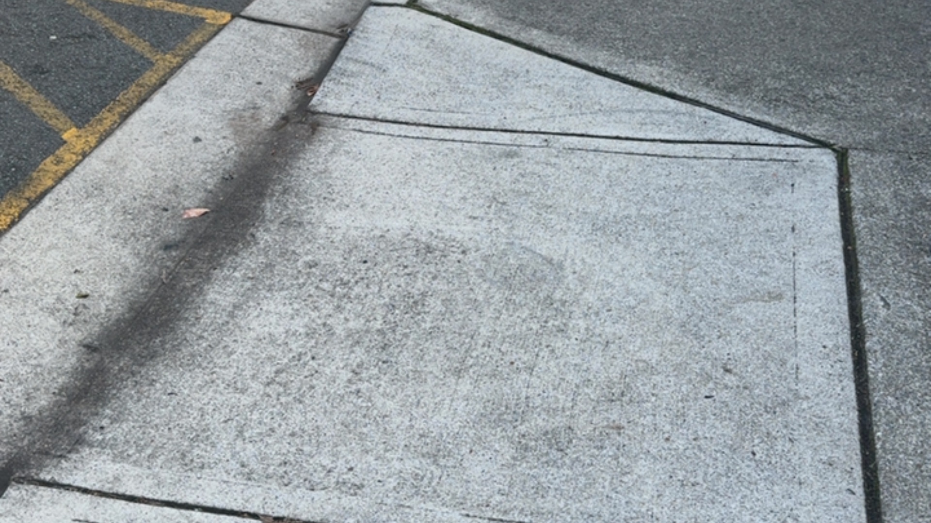 Textured light grey concrete curb ramp with a gradual incline, transitioning from dark asphalt marked with yellow diagonal lines. The pathway is wide and clear of obstructions.