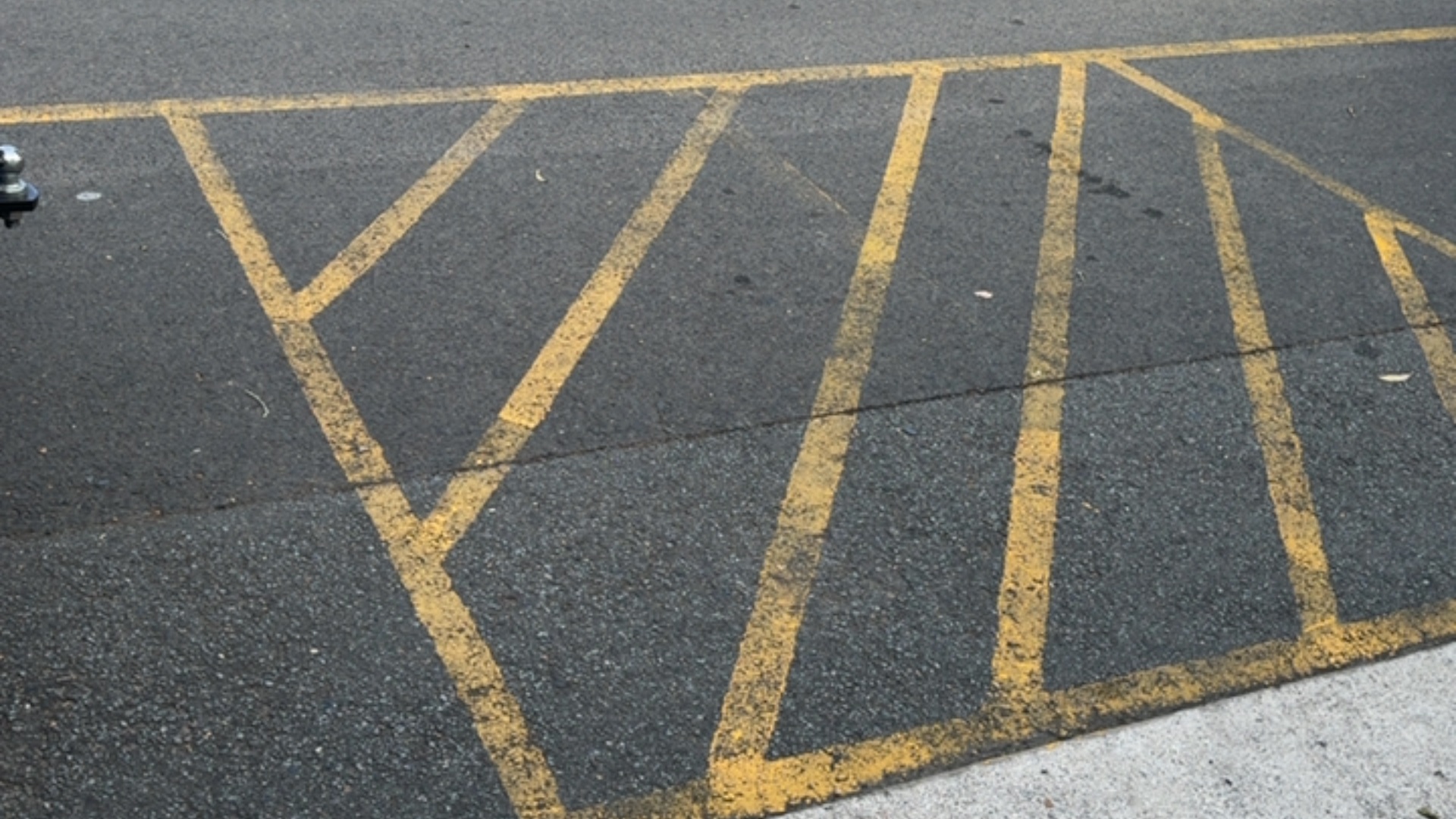 Yellow-painted hatched pattern on level dark grey asphalt, consisting of diagonal lines within parallel yellow borders, adjacent to a light-coloured concrete edge.