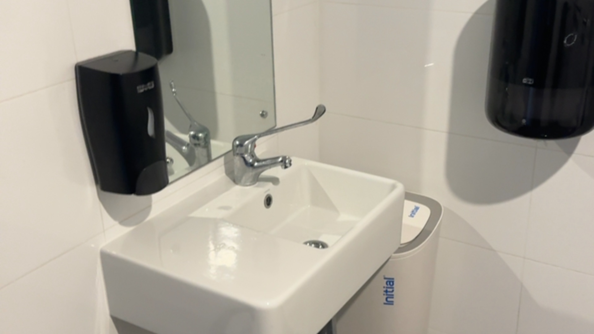 White wall-mounted sink with an extended lever tap. A soap dispenser and paper towel dispenser are mounted within reach on white tiled walls beside a hygiene bin.