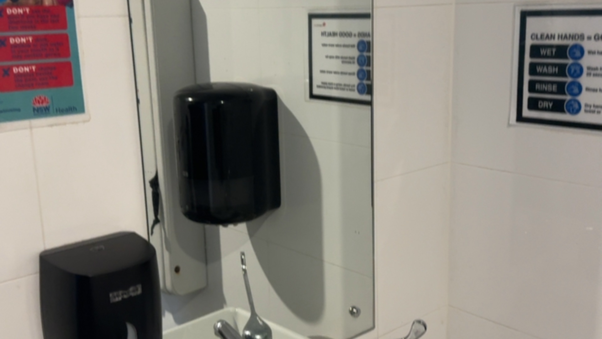 White tiled wall with sink, metal tap, and black wall-mounted dispensers. Mirror reflects signage alongside a 'CLEAN HANDS = GOOD HEALTH' guide and an NSW Health instructional poster.