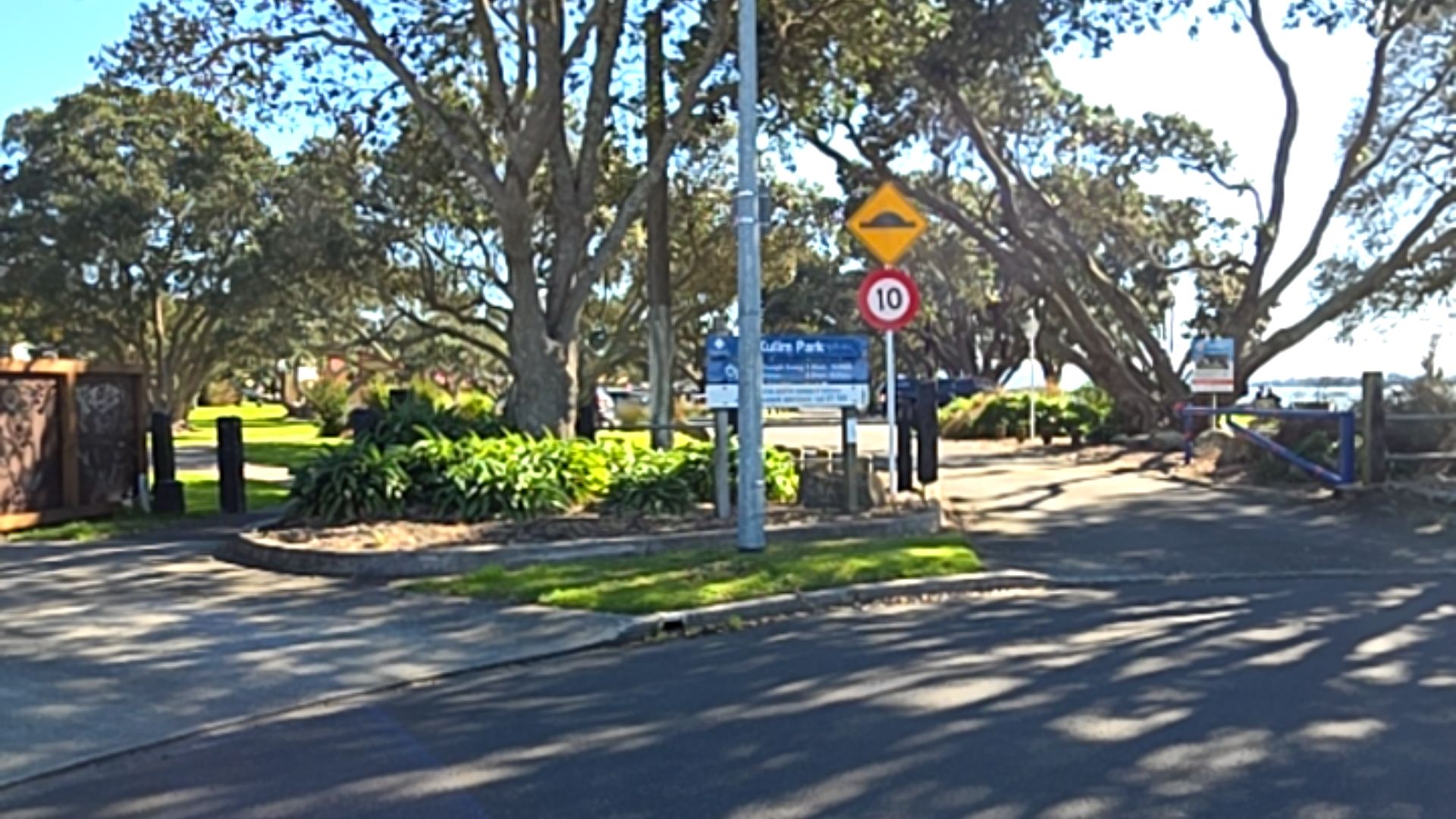 Level asphalt entrance to Kulim Park. Signs show a speed hump and 10 speed limit. Wide, shaded concrete pathways are bordered by mature trees, grass, and a blue metal gate.