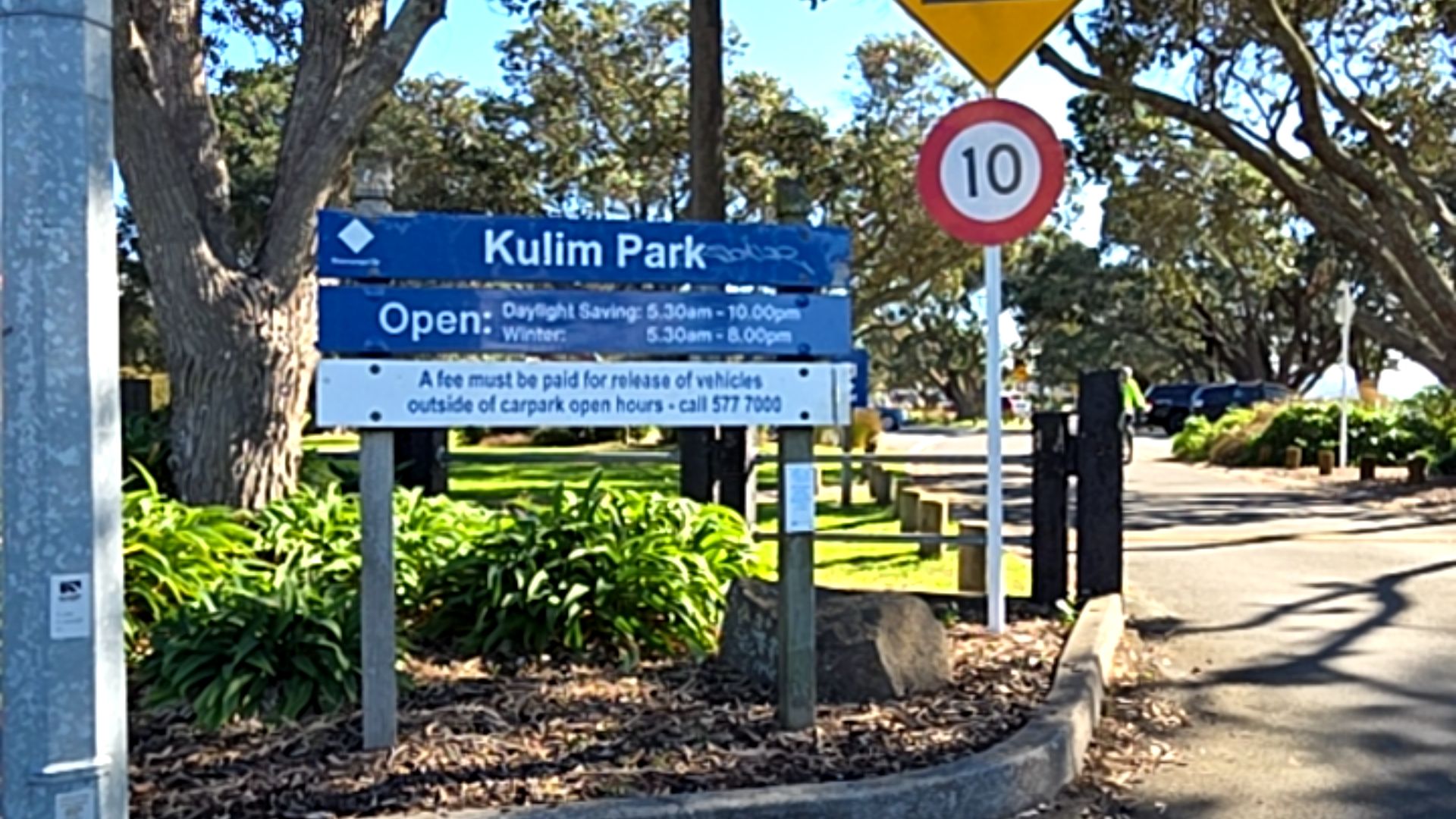 Level asphalt driveway entrance beside a blue Kulim Park sign listing opening hours and a red 10 speed sign. A low concrete kerb borders a garden bed with green foliage and timber signposts.