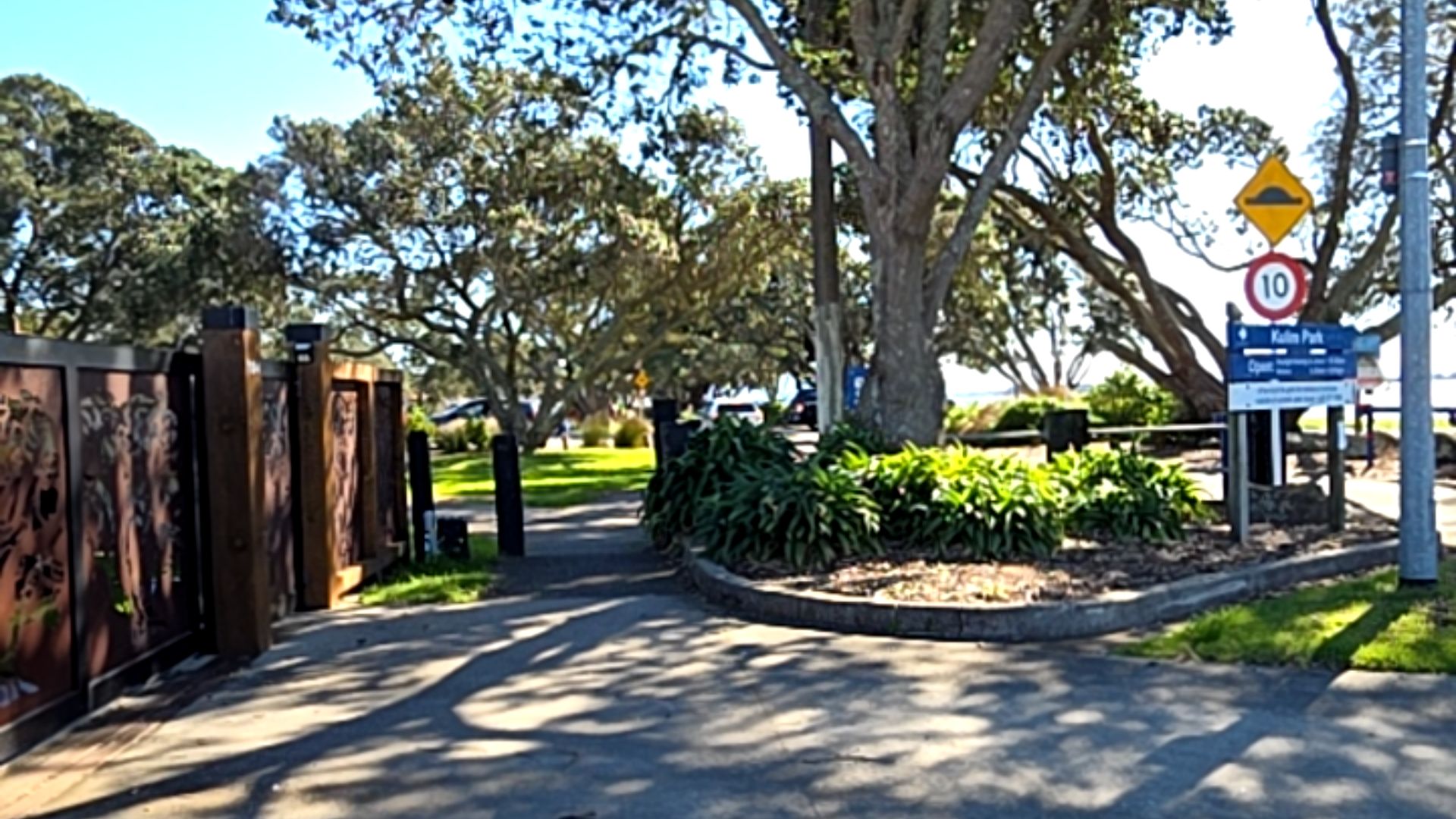 Wide level bitumen entrance at Kulim Park. Timber fence with decorative metal panels on the left. Signage indicates 10 speed limit. Large trees cast shade over the driveway and grassy area.