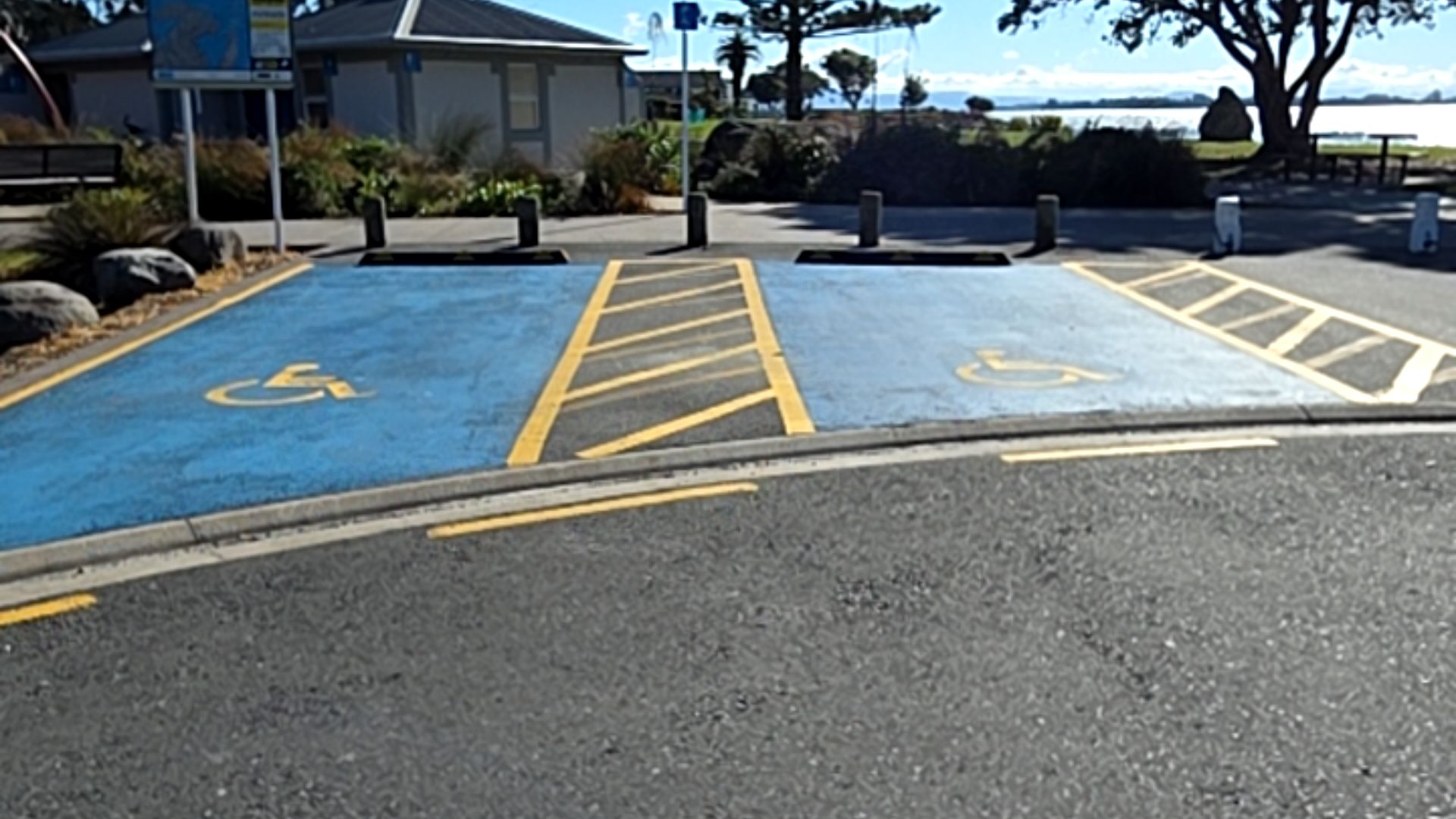 Two blue-painted accessible parking bays with yellow wheelchair symbols and hatched buffer zones. A sloped kerb ramp leads from level asphalt. Grey concrete wheel stops at the rear.
