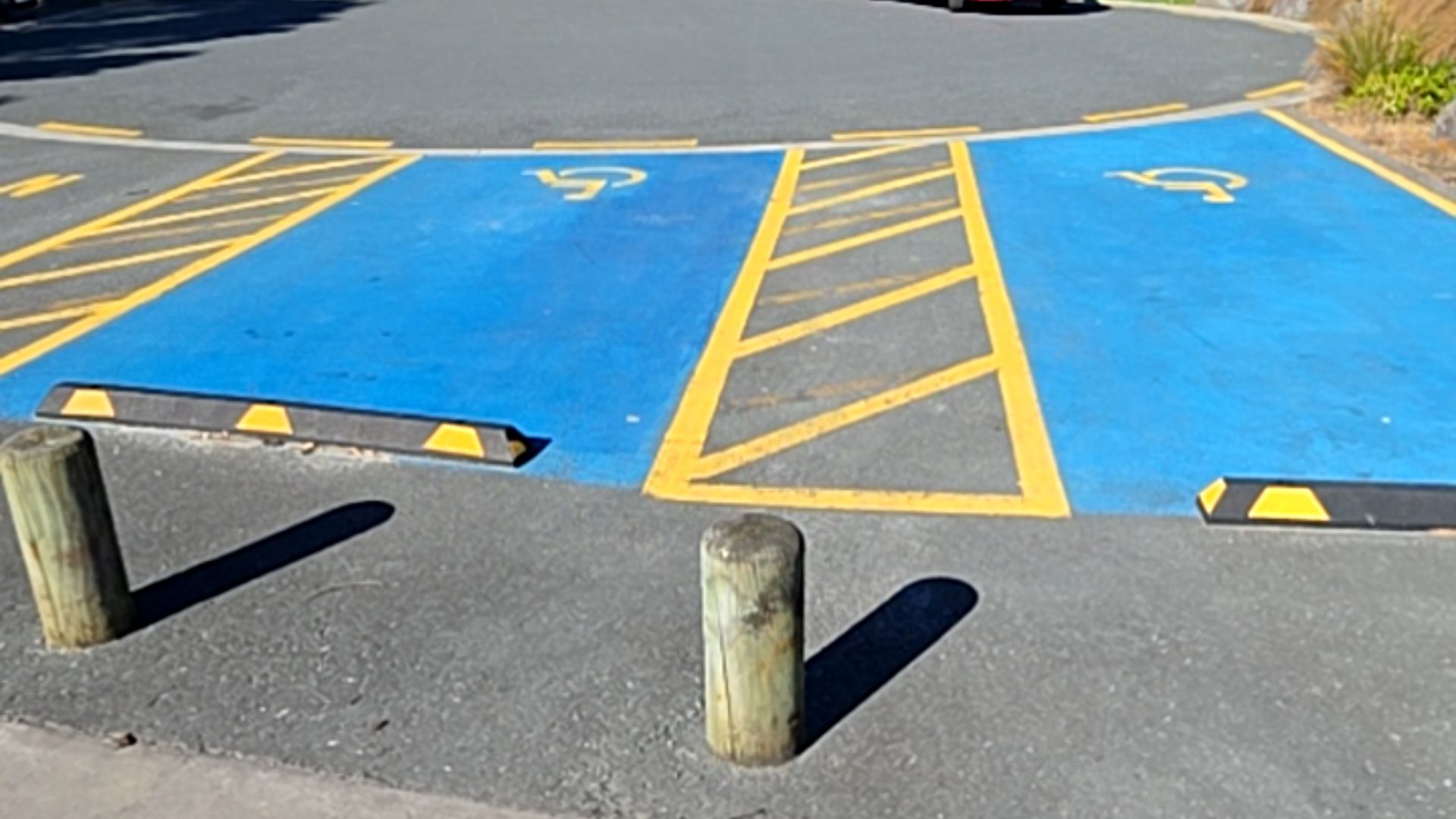 Two accessible parking spaces with blue ground paint and yellow wheelchair symbols. Yellow cross-hatched access zones sit between the spaces. Black wheel stops and wooden bollards are in front.