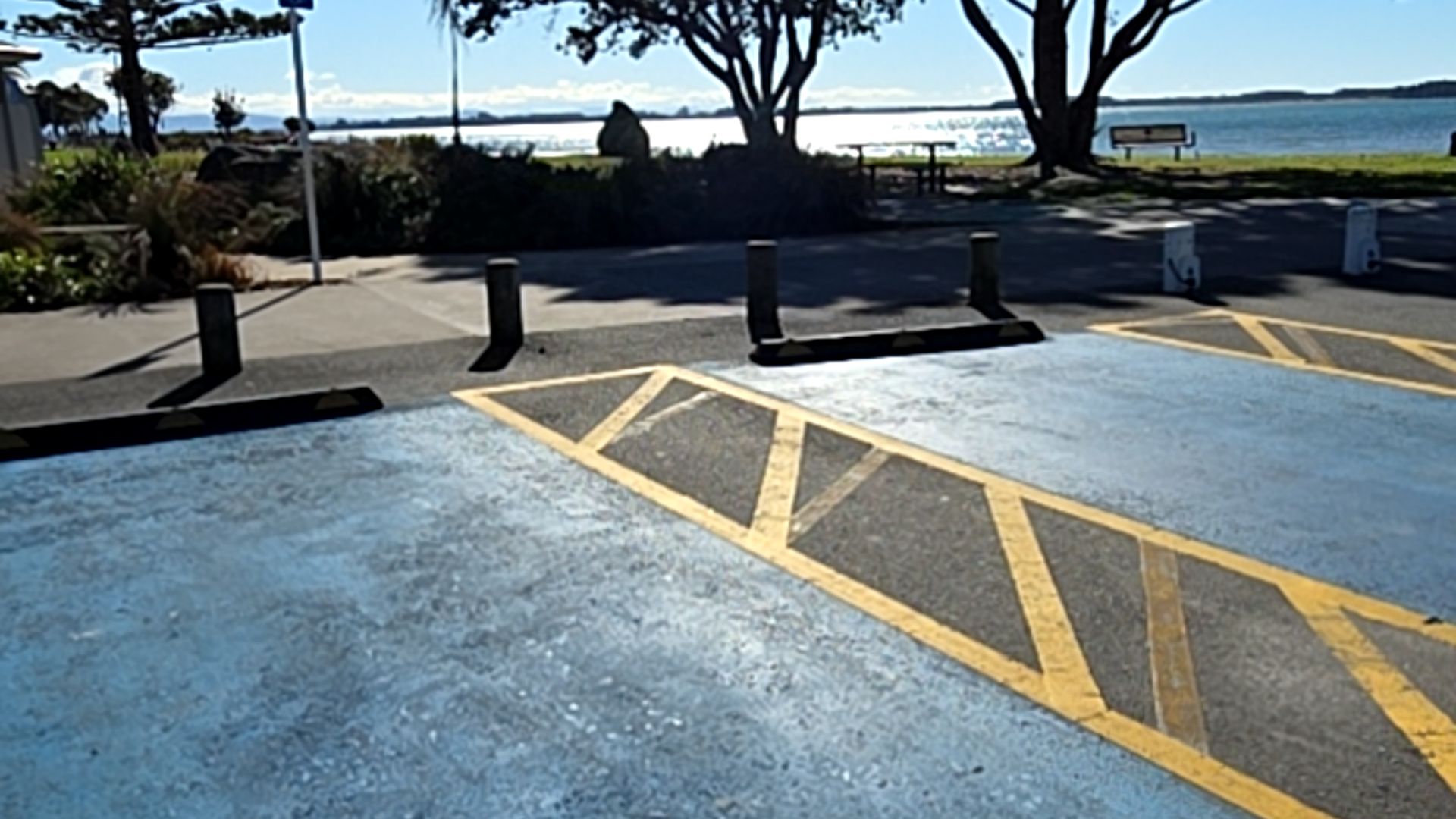 Blue-painted accessible parking bay with yellow hatch markings. Level concrete path behind grey bollards and wheel stops leads to parkland with picnic table, bench, and water views.