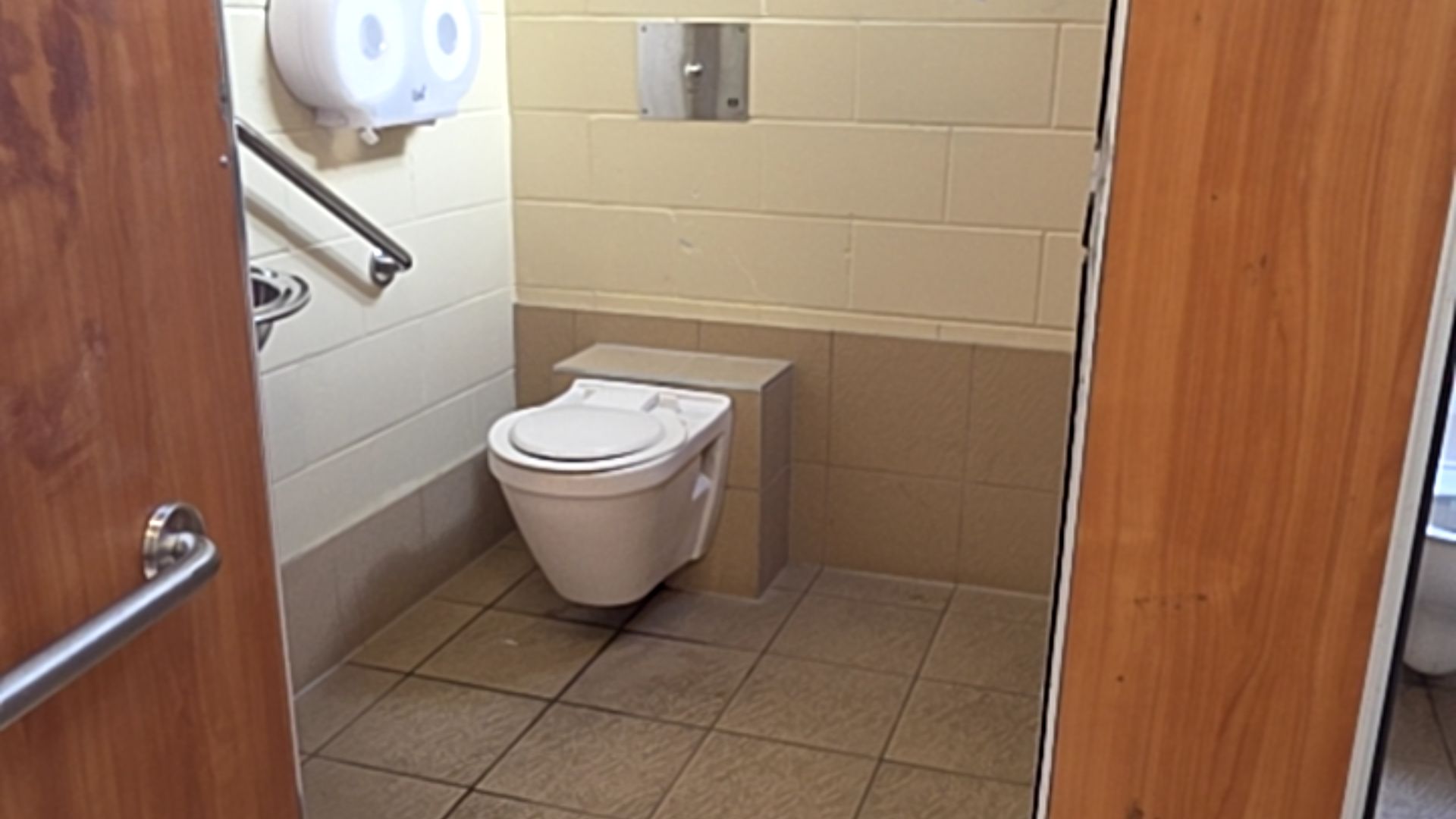 Wall-hung toilet with white lid. Diagonal metal grab bar on cream tiled wall below a white dispenser. Level beige tiled floor. Flush plate on wall. Wooden door with silver lever handle on left.
