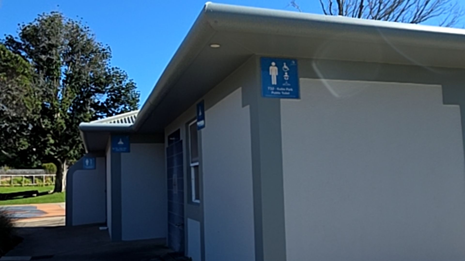 Public toilet block with blue signs showing male and wheelchair symbols. Recessed entrances are accessed via a level concrete path. Signage reads '710 - Kulim Park Public Toilet'.