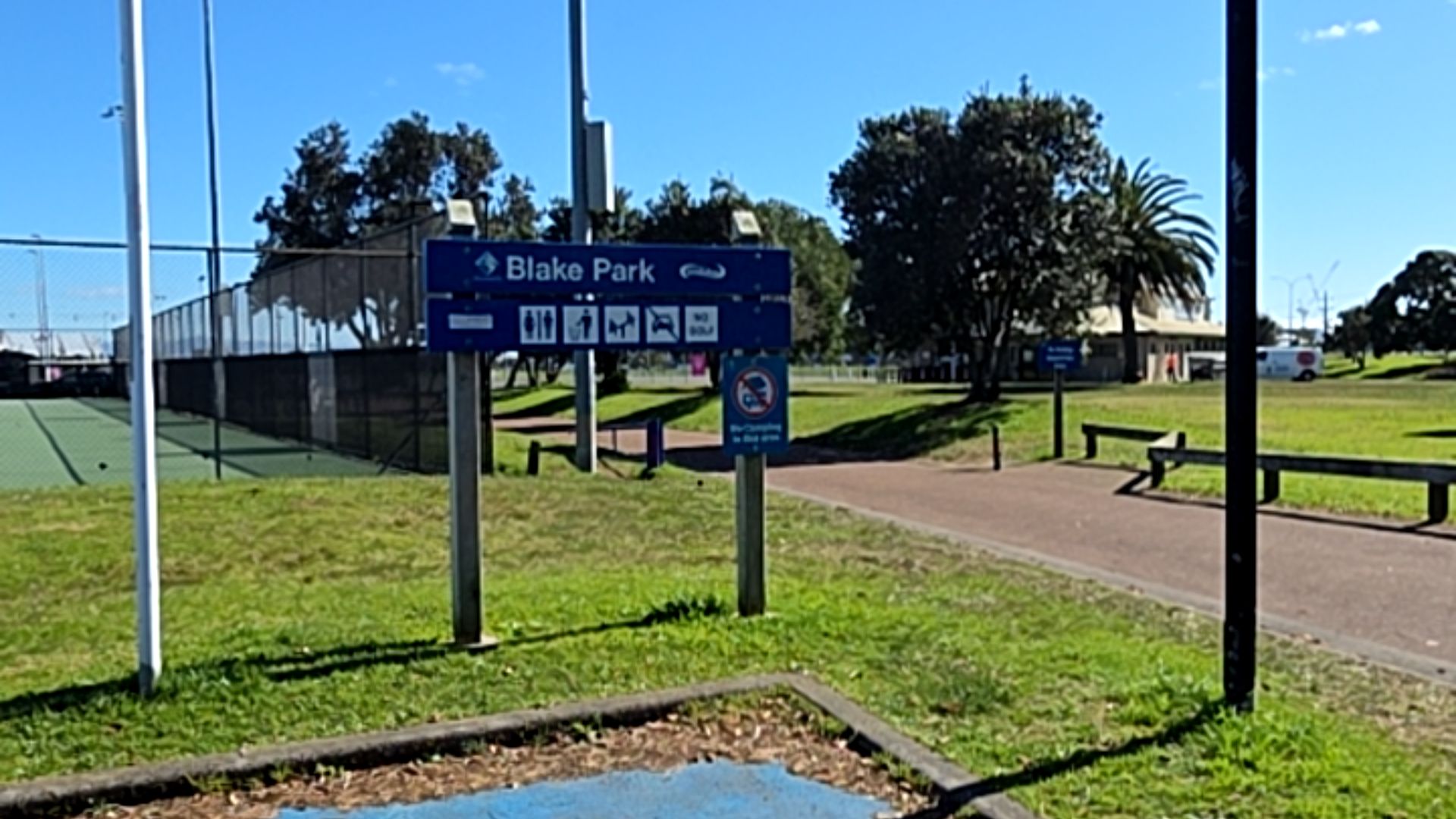 Level bitumen path past a blue Blake Park sign with icons for toilets, bins, and dogs on leash. Fenced tennis courts on the left with open grass and timber benches ahead.