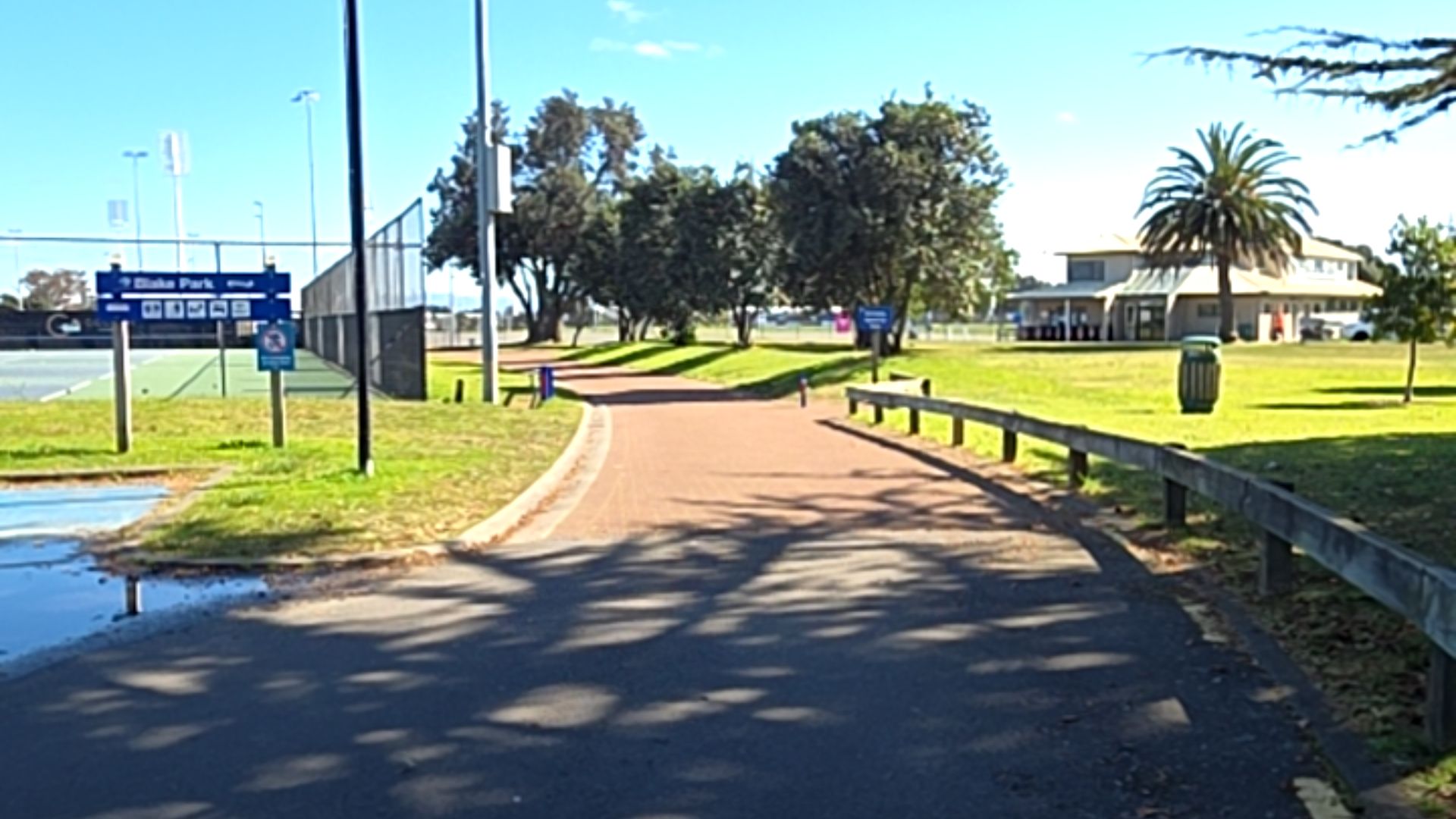 Wide, level bitumen and reddish-brown paved path curves right. Blue park signage on left near tennis courts. Timber post-and-rail fence borders grass on the right leading towards a building.
