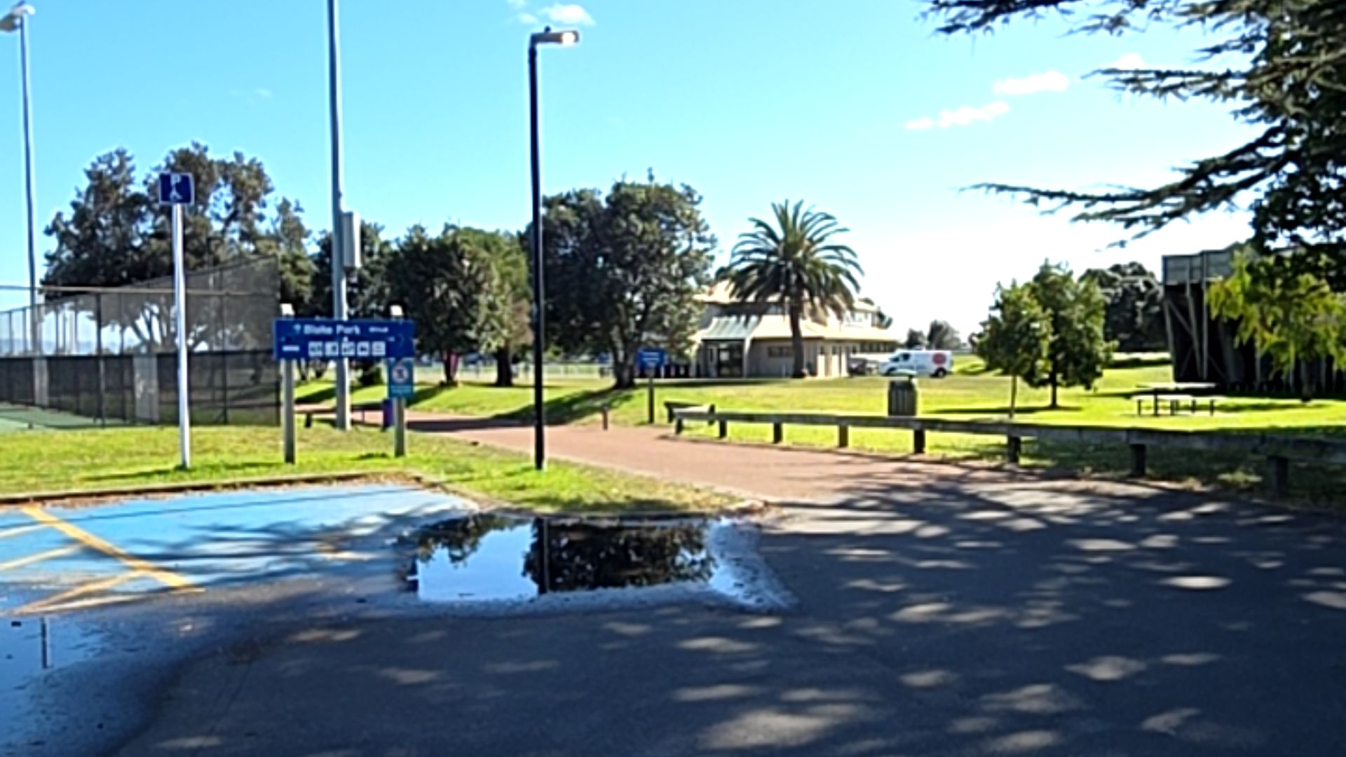 Blue accessible parking bay with white symbol beside a level reddish-brown path. Blue sign for Blake Park with icons. Path leads past a low timber rail towards a building. Puddle in foreground.
