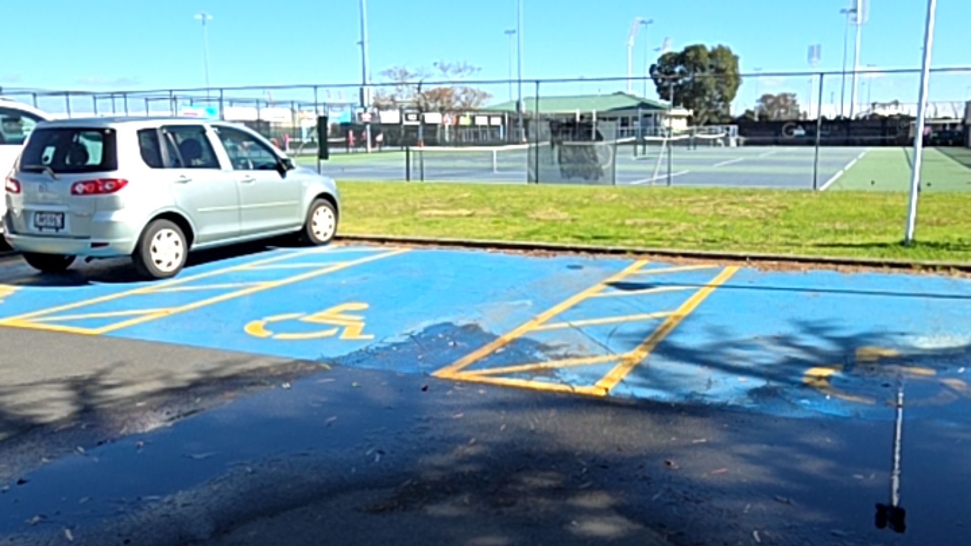 Two level blue accessible parking bays with yellow wheelchair symbols and shared hashed zone. Asphalt surface is wet with puddles. A silver car is parked in the left bay.