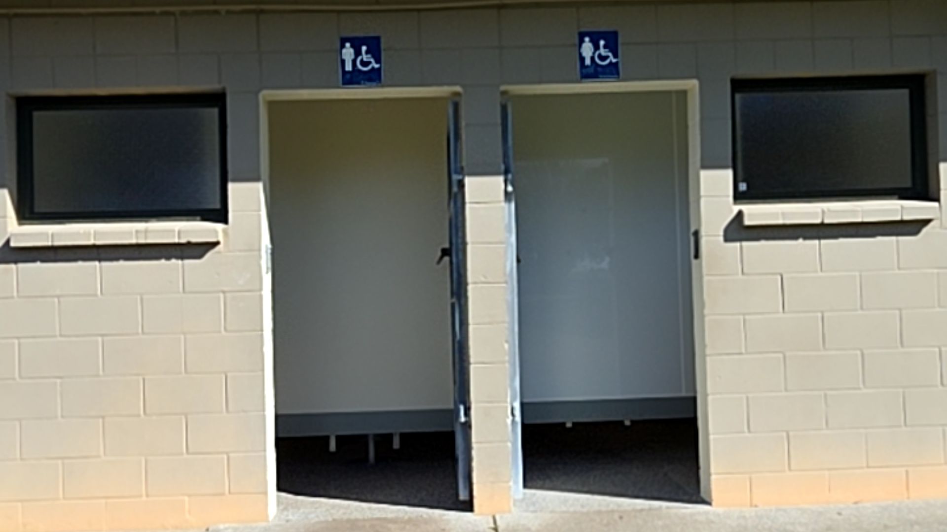 Two wide, level-entry doorways in a light-coloured concrete block wall. Blue signage with white male, female, and wheelchair accessibility symbols is above each entrance.