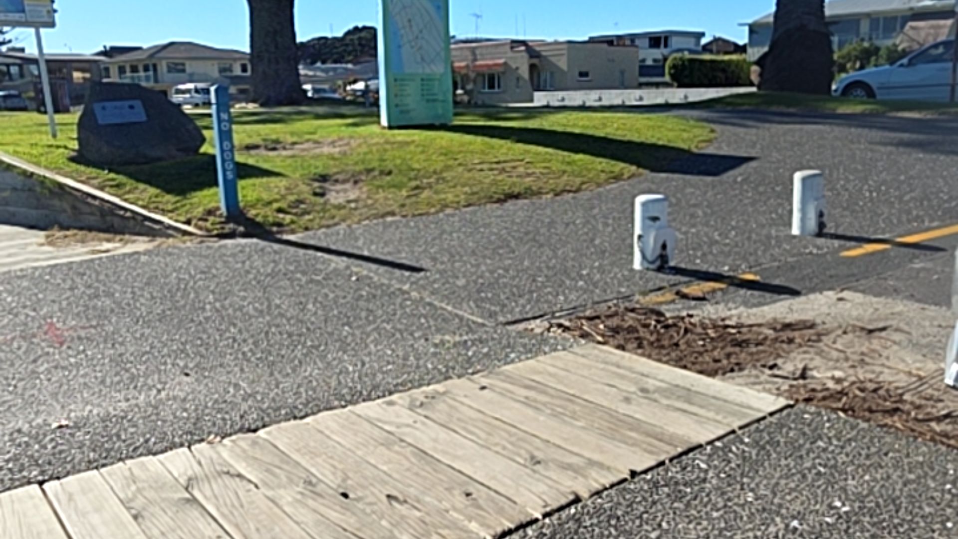 Wide, level asphalt path leads from a timber boardwalk. Two white bollards stand on the right. A blue 'NO DOGS' sign post is on the left. The surface is firm and unobstructed.