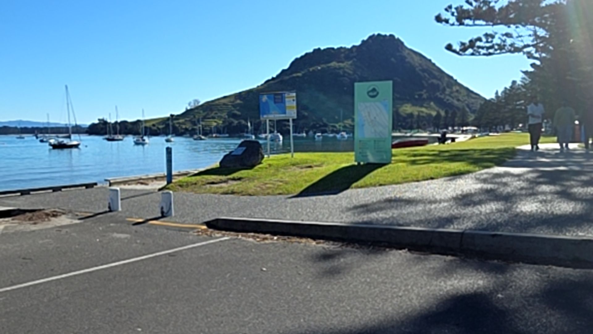 Level asphalt car park with low concrete kerbs meets a wide, flat paved pathway. Tall signage stands on a grass verge. A calm bay with yachts and a large hill are in the background.