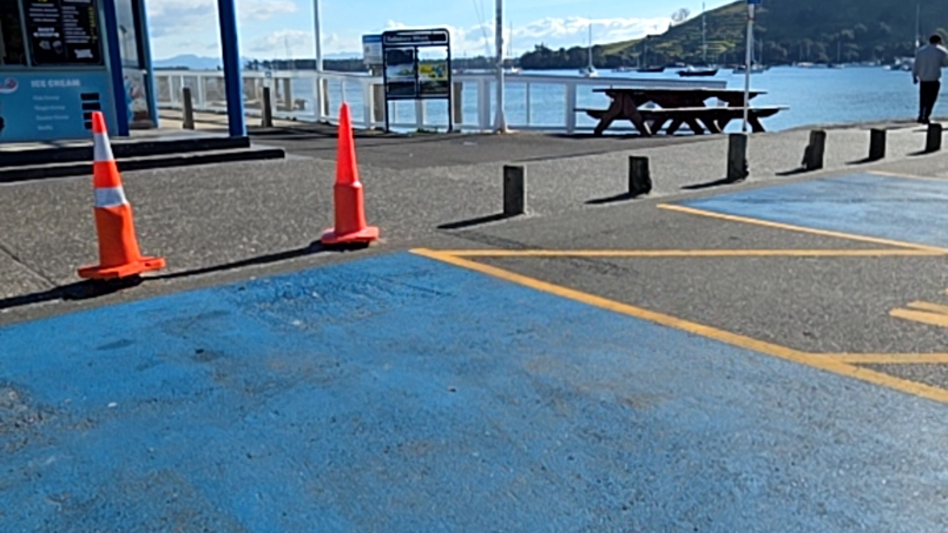 Level asphalt with blue and yellow paint markings and orange cones. Low bollards border a flat pedestrian area leading to a white timber-railed wharf, information sign, and picnic table.