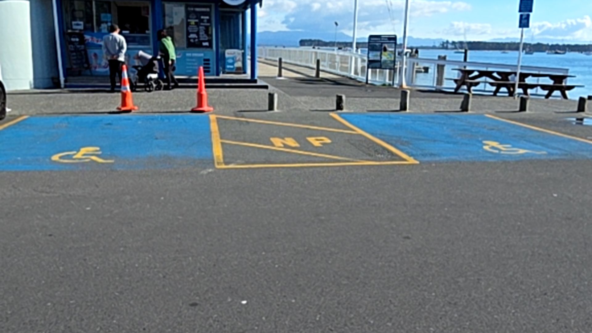 Two blue accessible parking bays with yellow symbols, separated by a yellow-lined zone marked 'NP'. Flat asphalt surface with orange cones near a shop with a level entrance.
