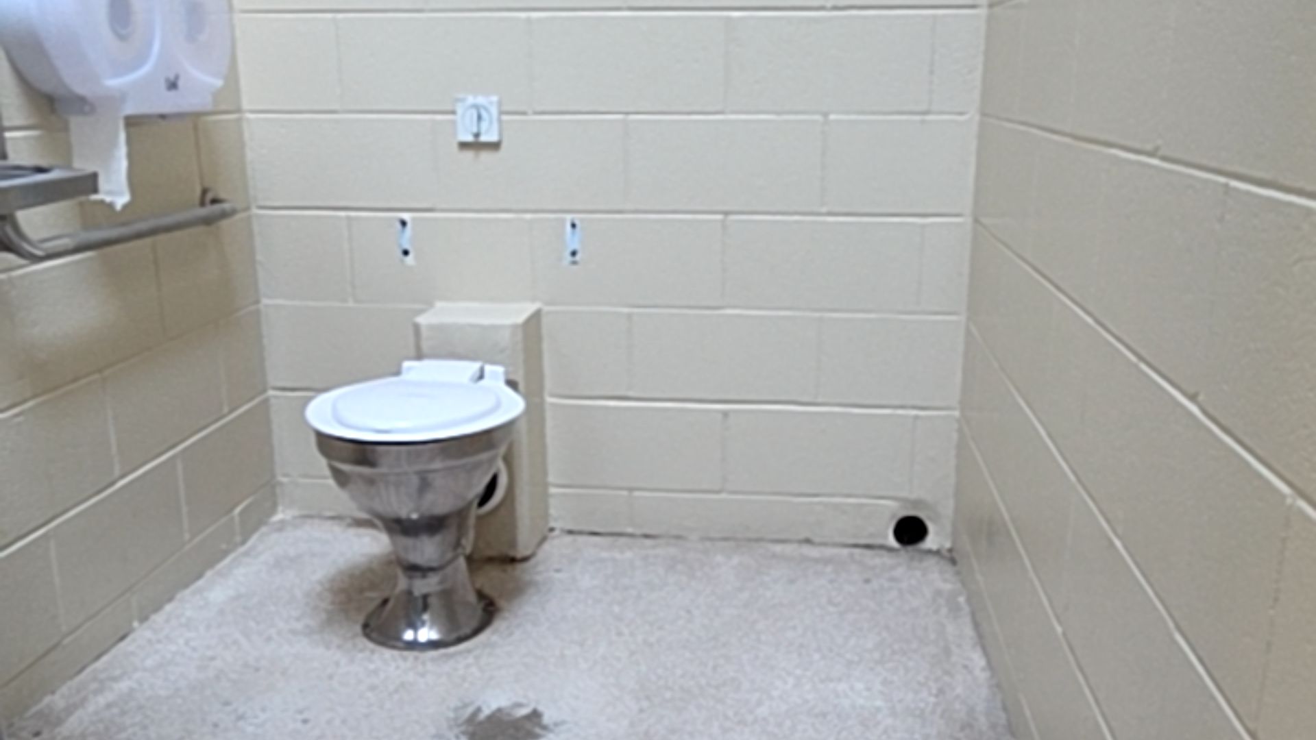 Stainless steel toilet with white lid. Beige masonry walls and light grey concrete floor. Horizontal stainless steel grab rail on left wall beside a white toilet paper dispenser.
