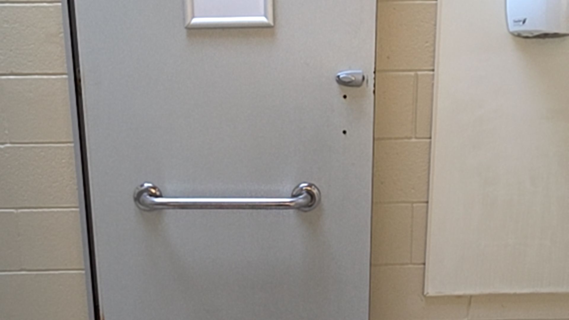 Light grey door with a horizontal silver metal grab bar and a thumb-turn lock. Set against a beige brick wall with a white panel and hand dryer visible to the right.