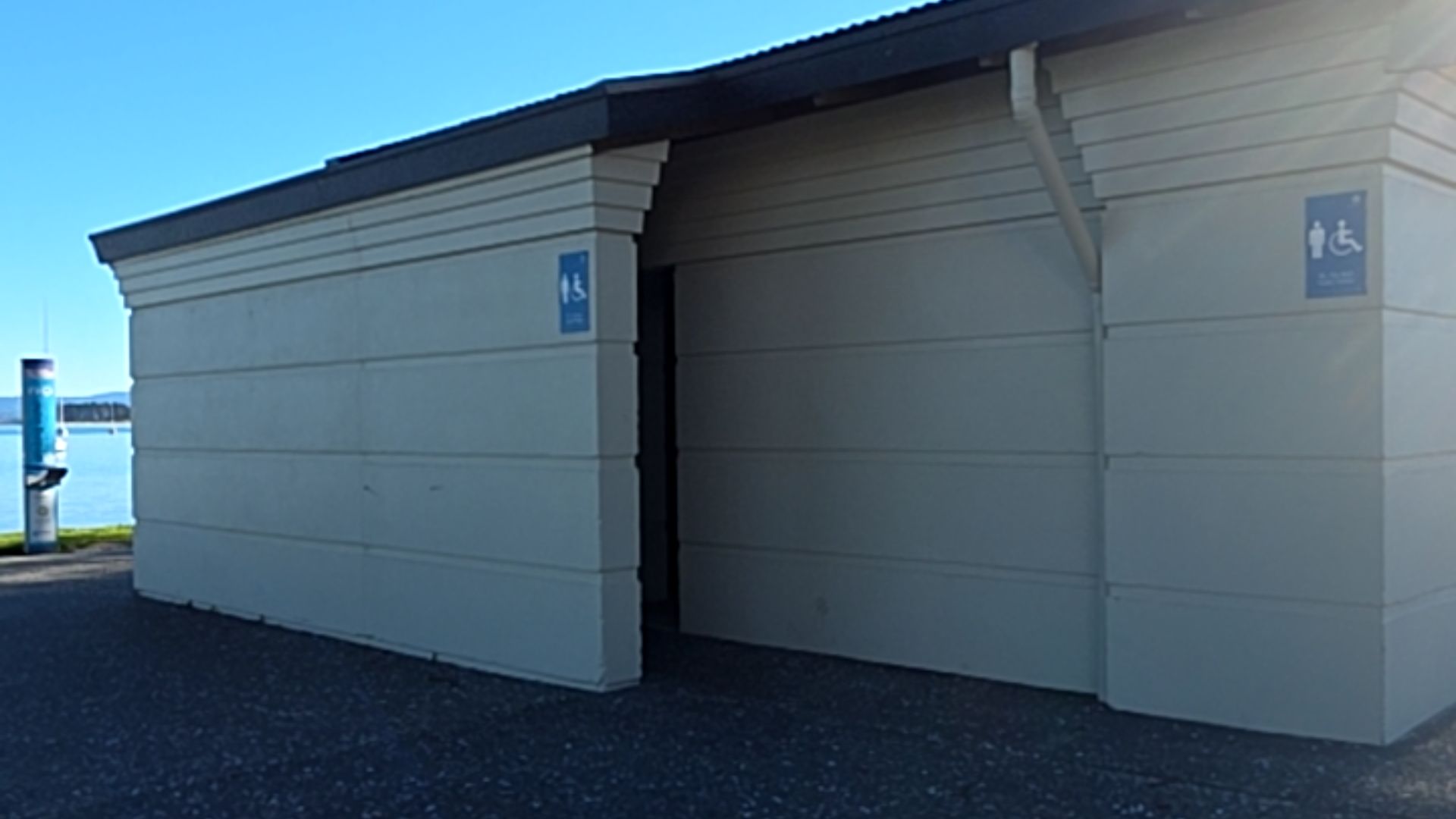 Level dark grey pavement leads to an open entrance in a beige building. Two blue signs on the exterior walls display male and wheelchair accessibility symbols.