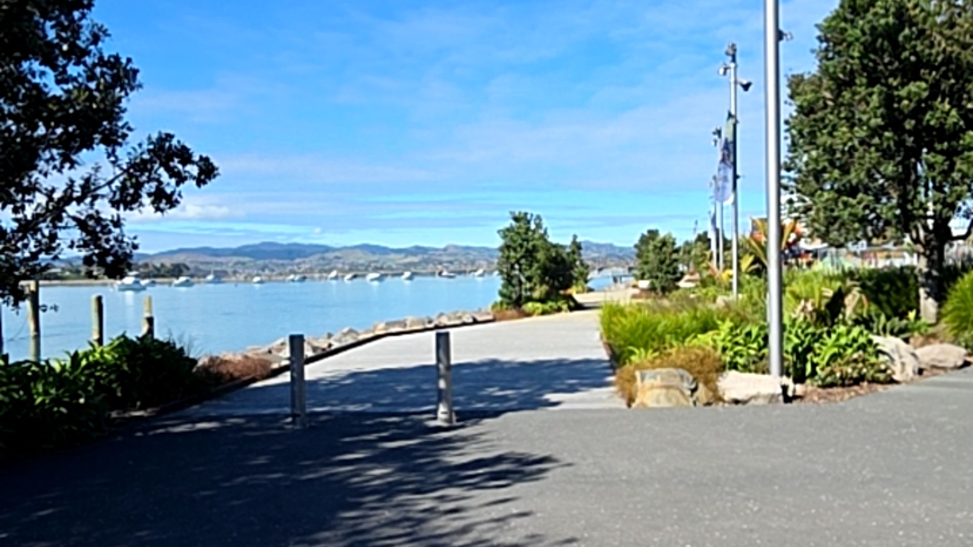 Wide, level concrete pathway along a waterfront. Two silver metal bollards mark the entrance from a smooth asphalt surface. Lush greenery and large rocks line the side of the flat path.