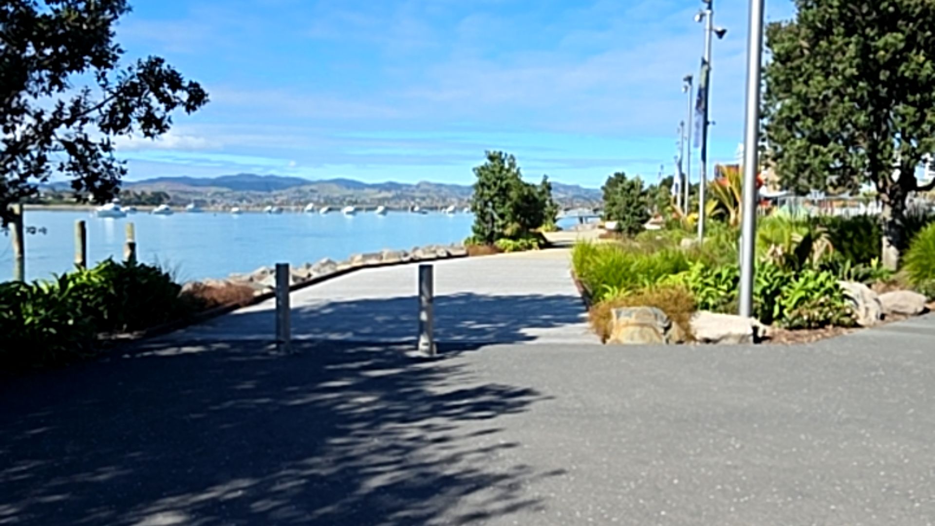 Wide, level asphalt and concrete pathway with two vertical timber bollards. The flat surface is bordered by low garden beds and tall metal poles, leading along a harbour.