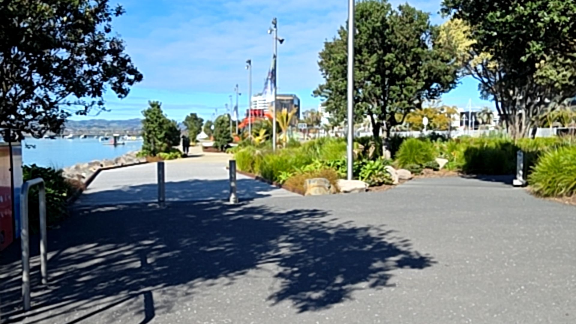 Wide, level asphalt and concrete pathway with grey metal bollards and a handrail. Smooth surface runs alongside water with lush green landscaping. No visible stairs or obstructions.