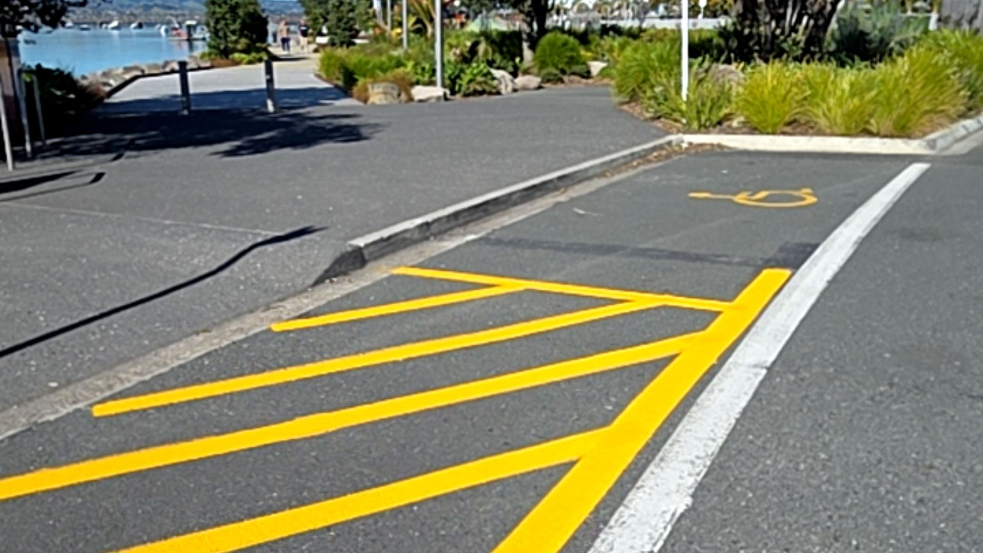 Accessible parking bay with yellow wheelchair symbol and hatched shared zone. A concrete kerb ramp leads to a wide, level asphalt pathway.