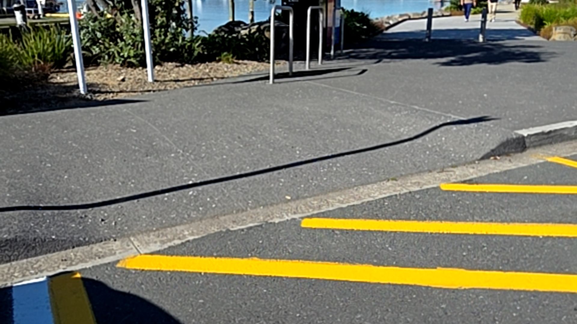 Level concrete kerb ramp connecting an asphalt carpark with yellow markings to a wide paved footpath. A thin black cable lies across the path near the ramp transition.