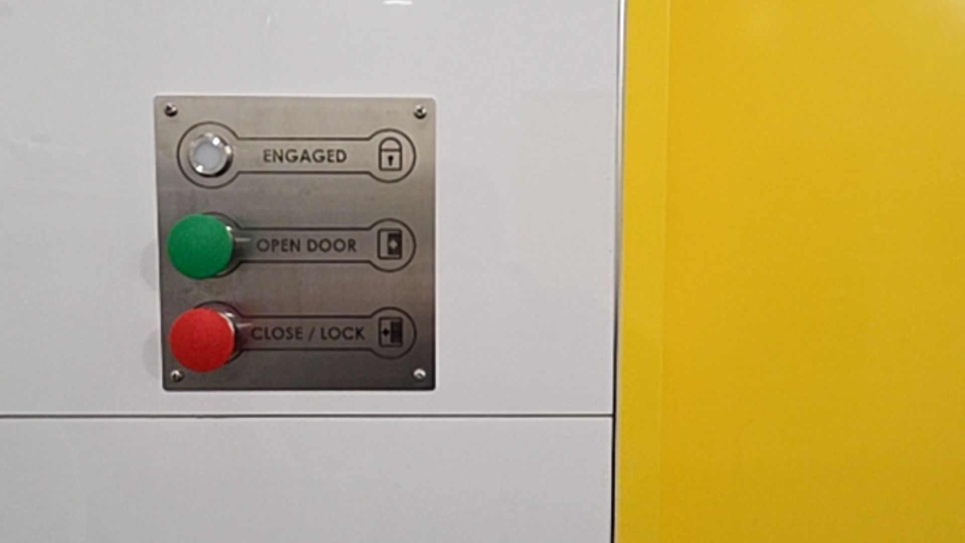 Stainless steel door control panel on white wall. Green OPEN DOOR button, red CLOSE / LOCK button, and ENGAGED light indicator. Features large text and high-contrast icons for accessibility.