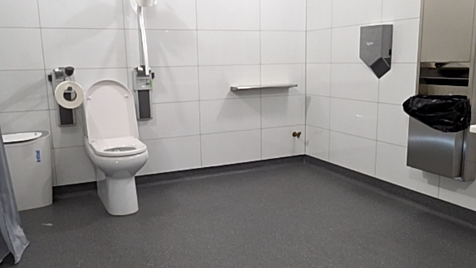 White toilet with vertical and horizontal grab rails on white tiled walls. Spacious dark grey non-slip floor. Wall-mounted grey hand dryer and recessed waste bin on the right wall.