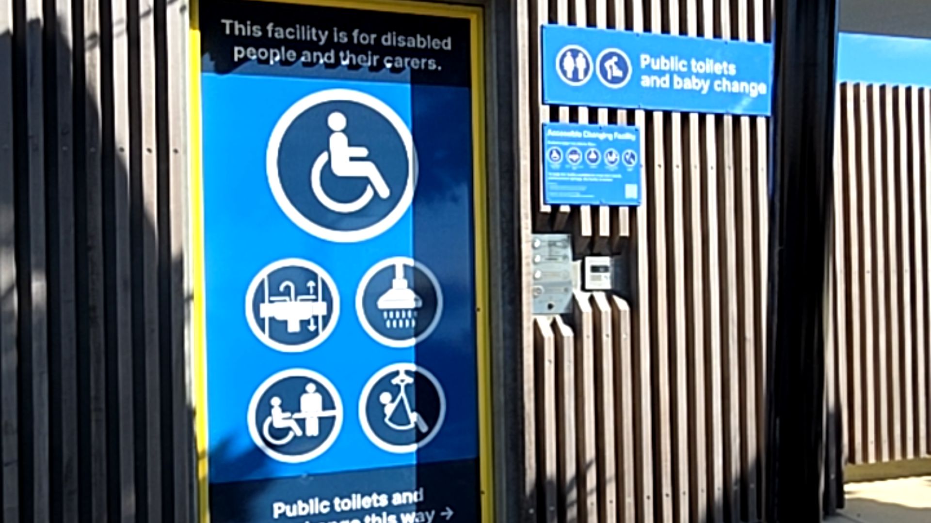 Blue sign on vertical timber slats with symbols for wheelchair, adjustable sink, shower, change table, and hoist. Signs on the right point to public toilets and baby change facility.