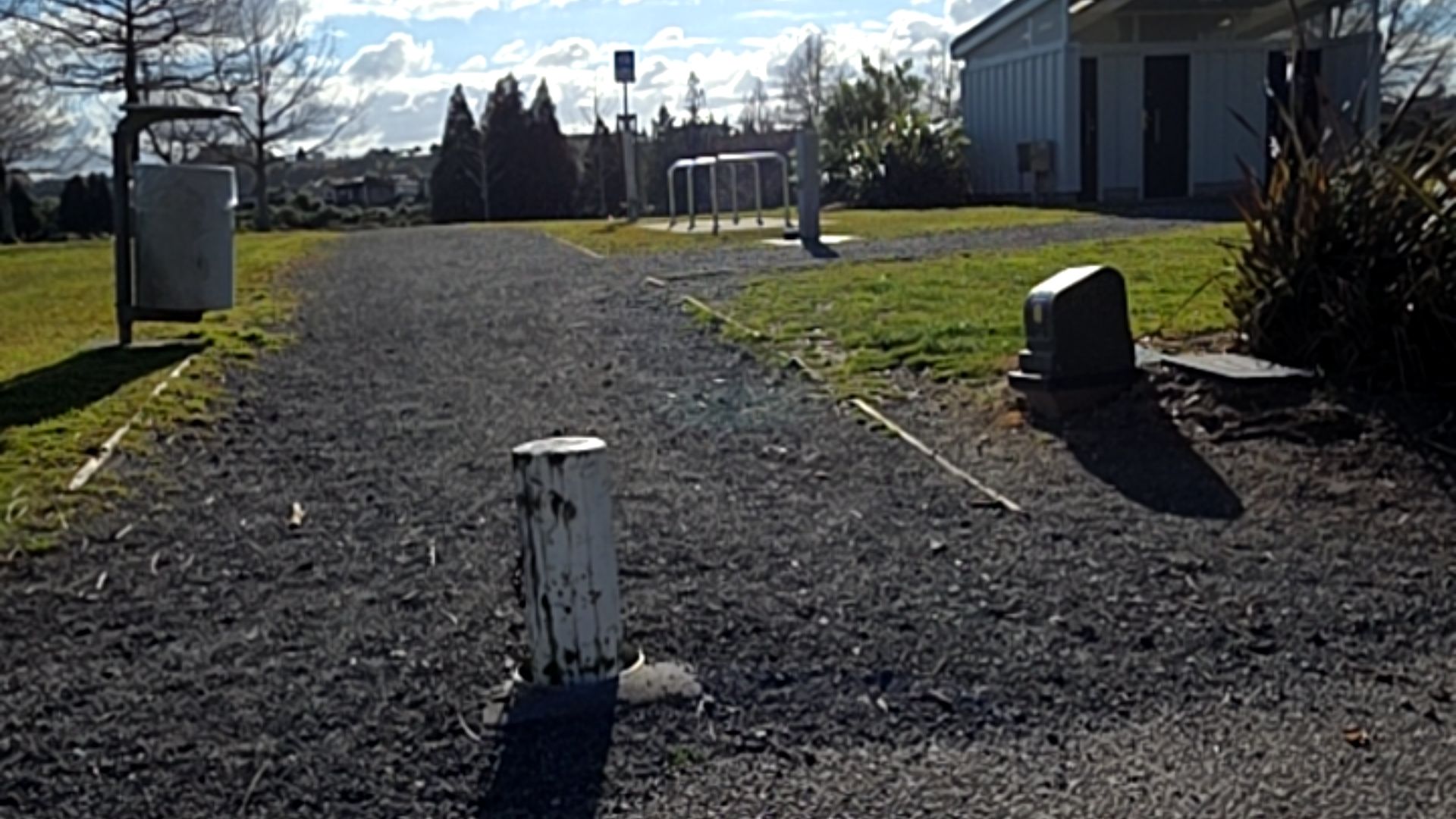 Level gravel path with a central white bollard. A silver rubbish bin is on the left. In the background are metal bike racks on concrete and a grey public building with level access.