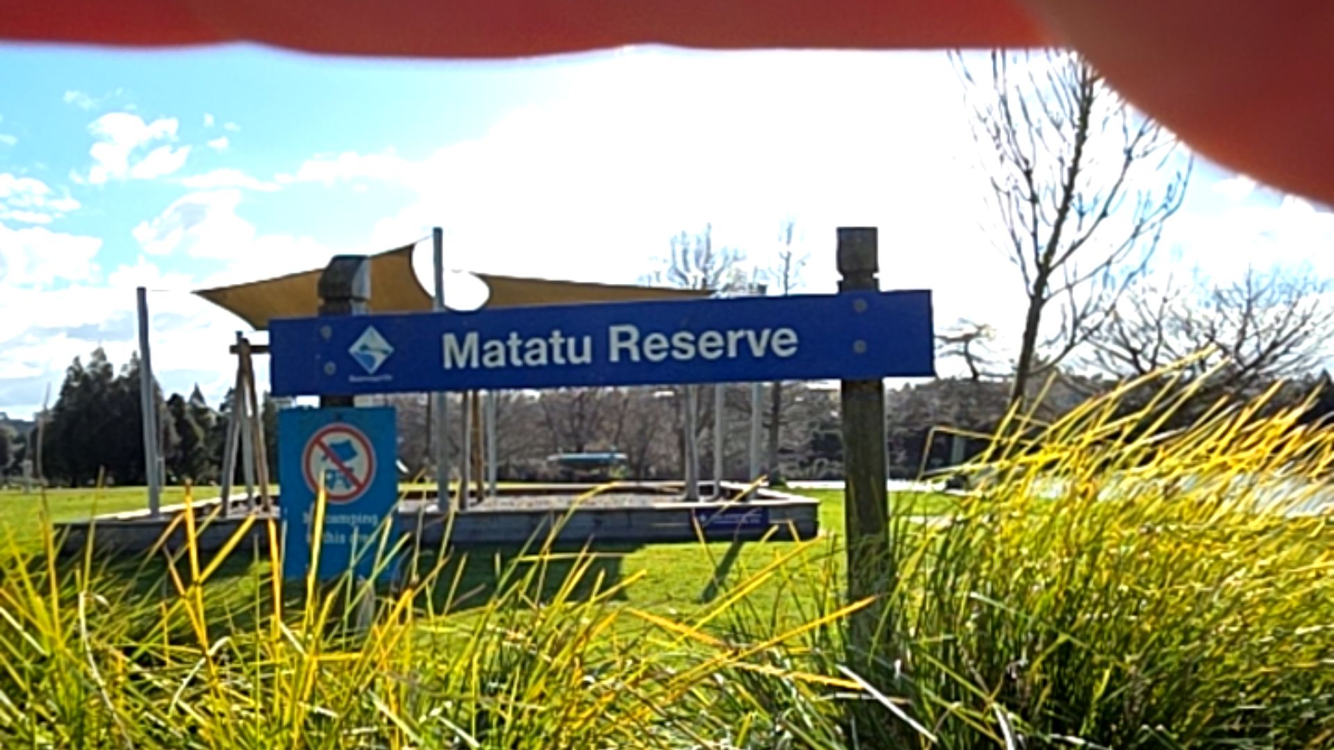 Horizontal blue sign reading 'Matatu Reserve' on timber posts. Smaller 'no camping' sign below. Level grass leads to a timber-edged playground area under a tan shade sail.