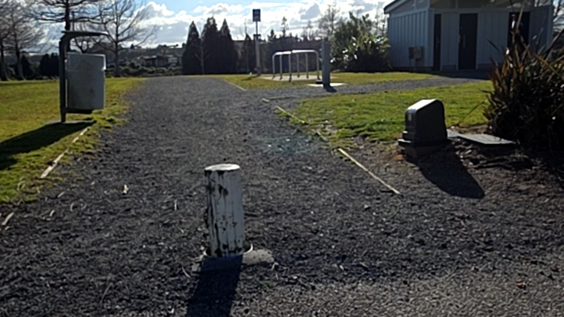 Wide level gravel pathway with a white bollard in the foreground and a silver rubbish bin to the left. Silver bike racks and a grey building are visible in the background.