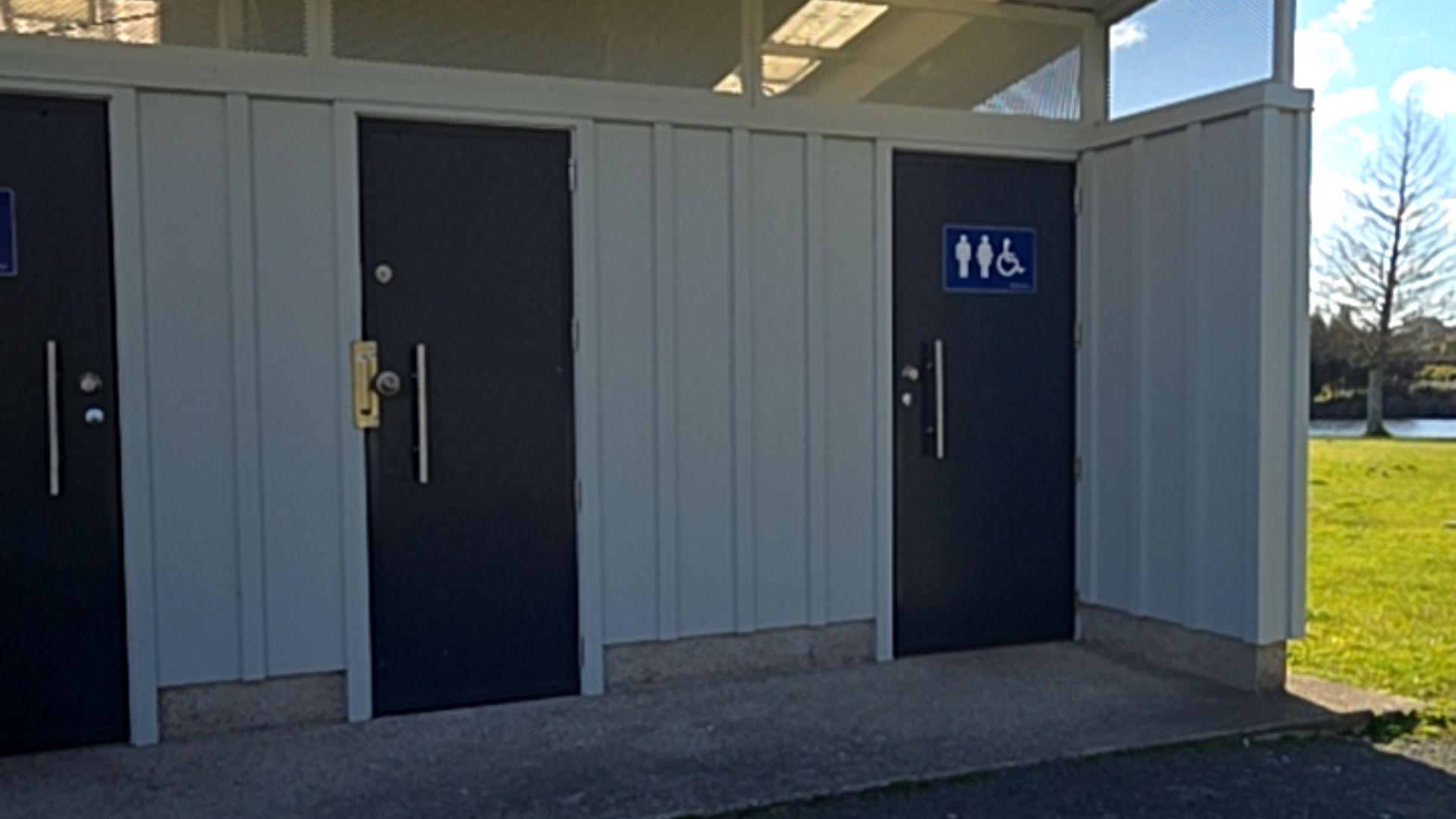 Three dark grey doors on a level concrete surface. Right door features a blue sign with white male, female, and wheelchair symbols. Doors have vertical silver pull handles and locks.