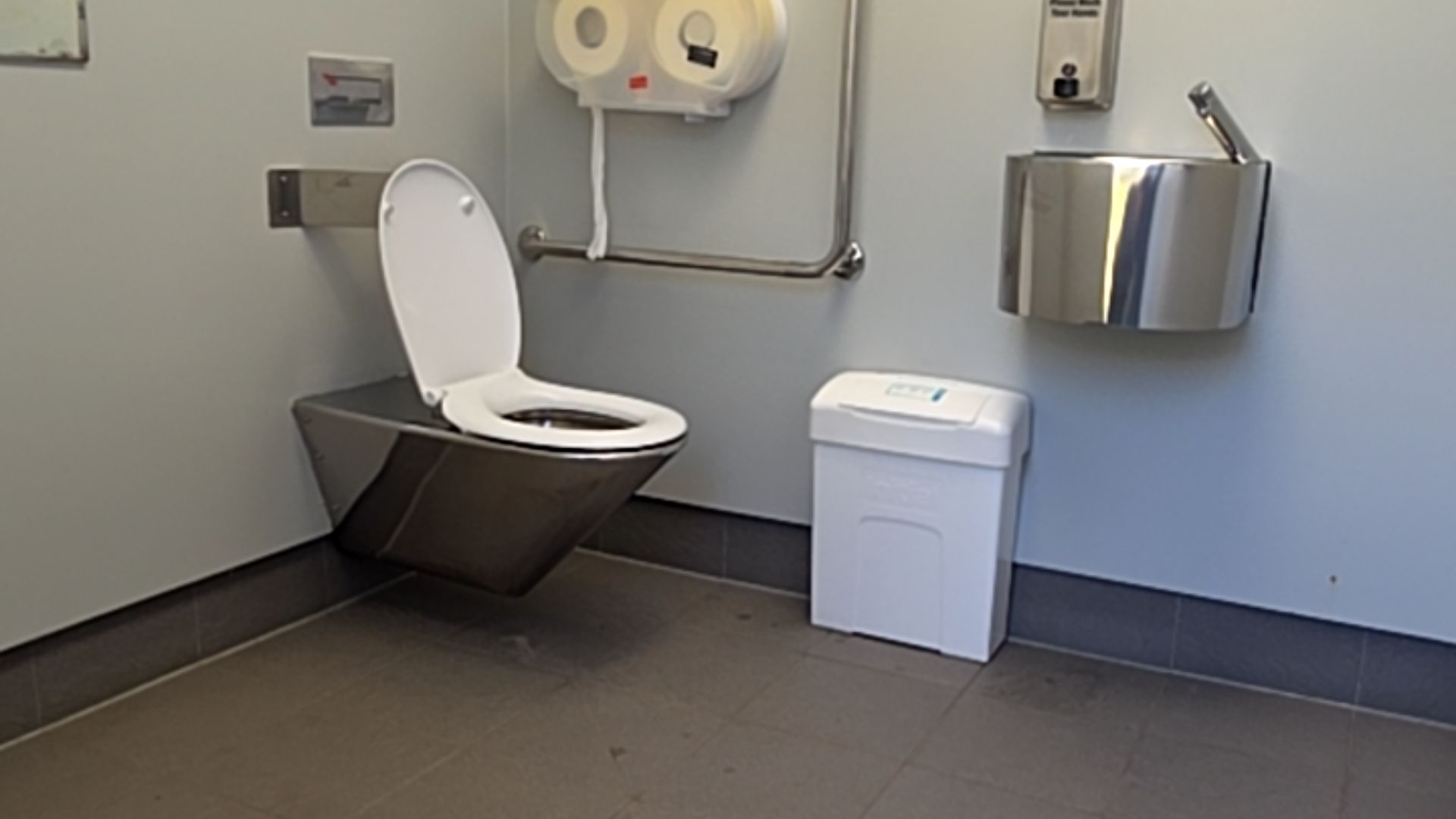 Wall-mounted stainless steel toilet with white seat and horizontal grab rail. Wall-mounted sink and white bin on dark grey tiled floor. Light grey walls with soap and toilet paper dispensers.