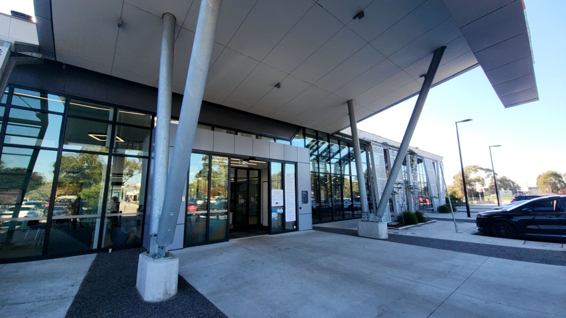 Level concrete entrance with automatic glass sliding doors under a sheltered canopy. Step-free pathway with V-shaped metal columns and signage on the glass. Bitumen car park to the right.