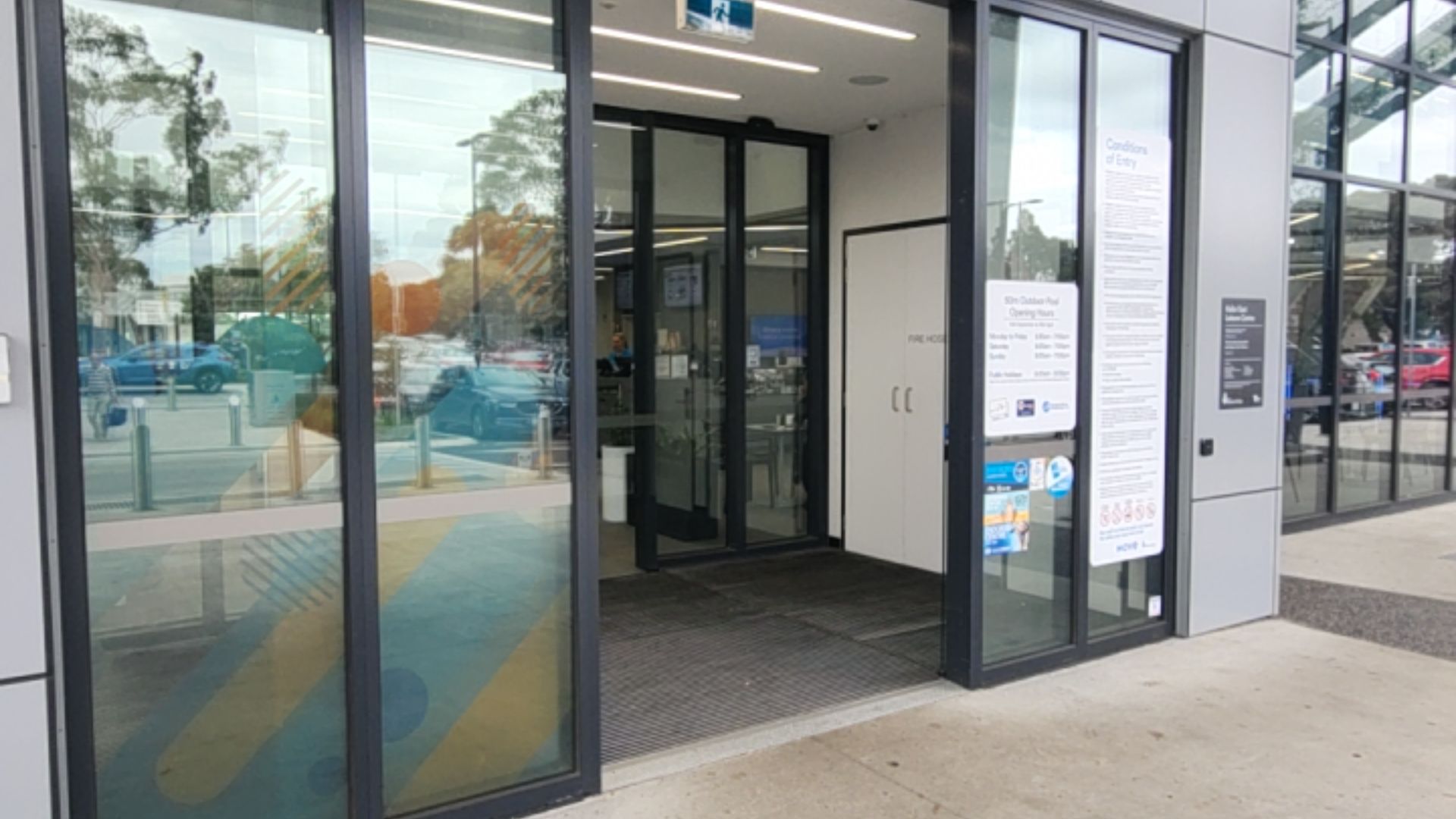 Wide level entrance with automatic glass sliding doors and dark grey textured floor mat. Light grey concrete pathway leads to the threshold. White signage is visible on the glass and wall.
