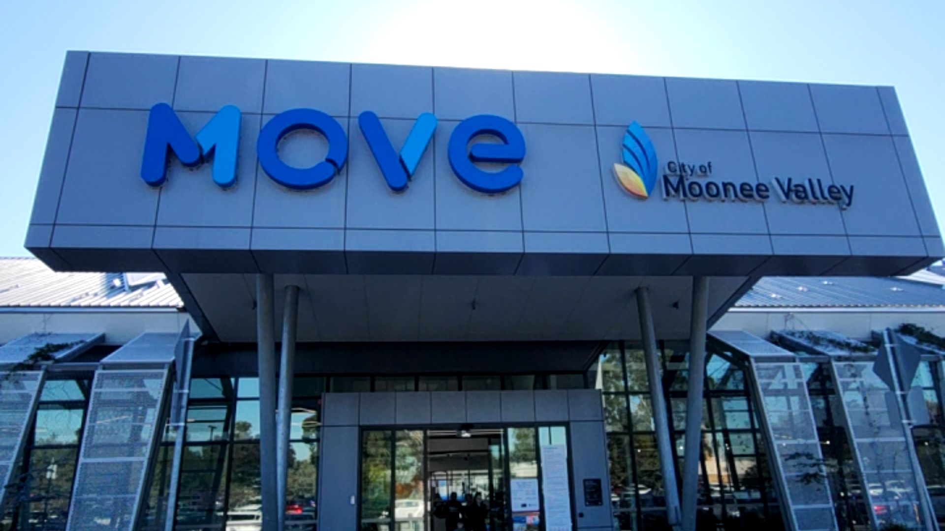 Level entrance with automatic glass sliding doors under a grey panelled facade. Large blue 'MOVE' and 'City of Moonee Valley' signage. Metal pillars support the overhanging roof.