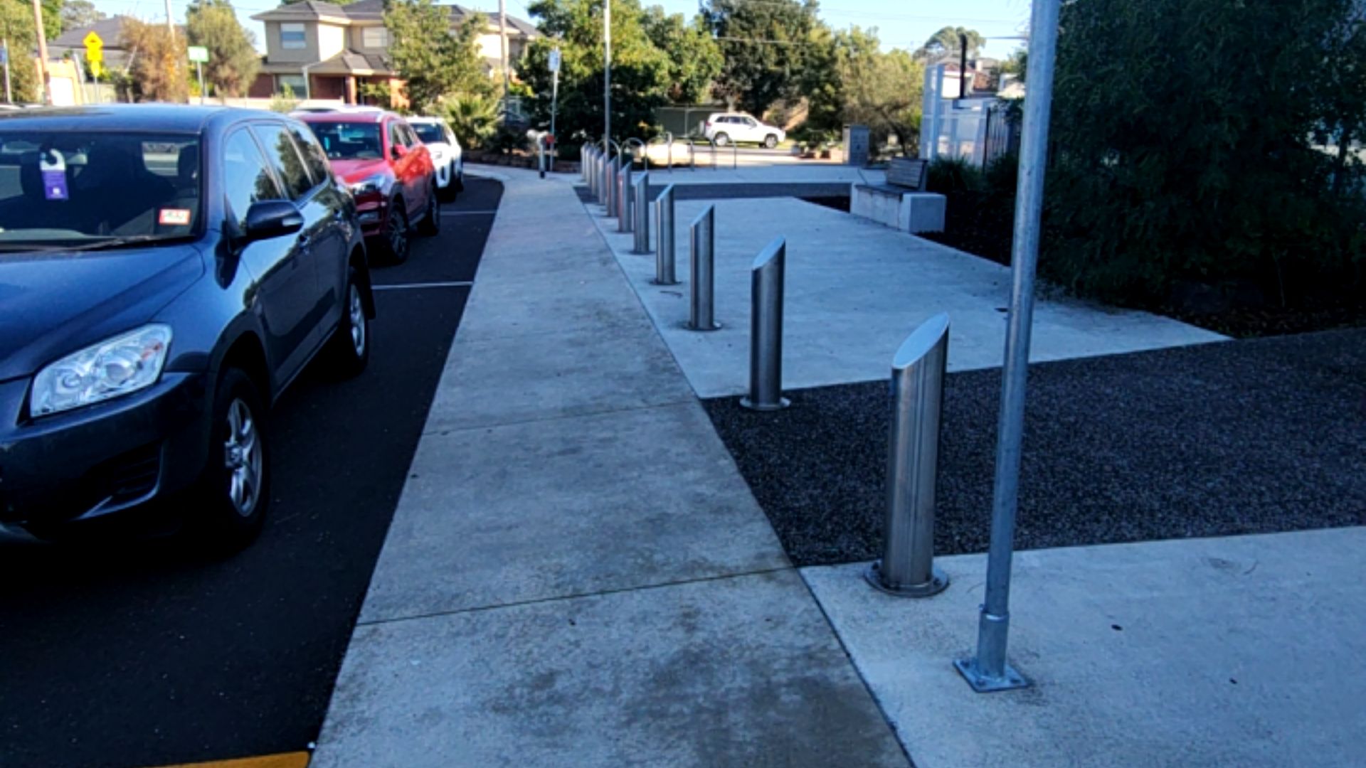 Wide, level concrete footpath bordered by slanted silver metal bollards. Adjacent parking bay with a dark grey car. Smooth pavement leads to a white bench seat in the background.