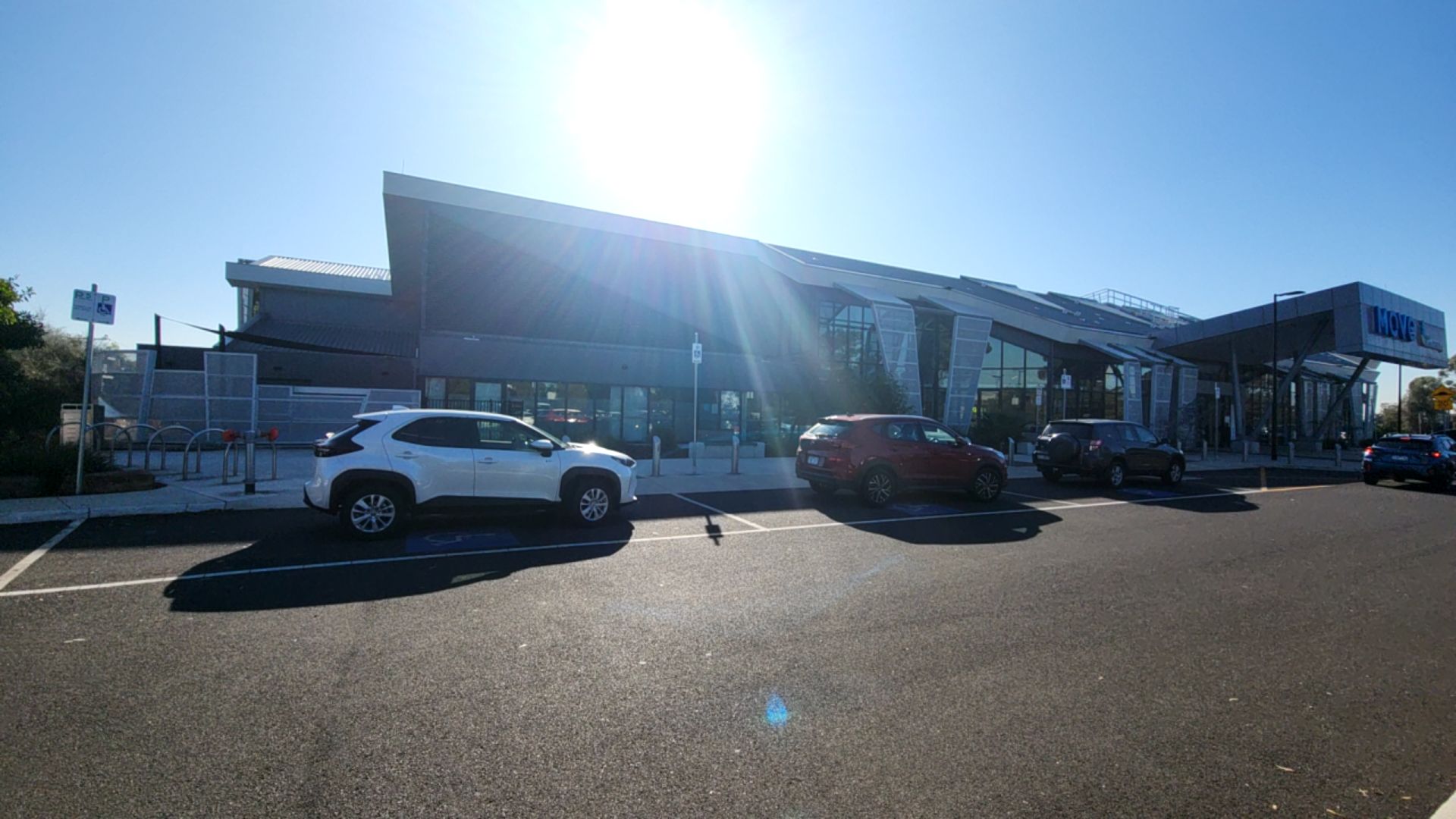 Two accessible parking spaces with blue wheelchair symbols and signage on level bitumen. A modern glass-fronted building with a wide, level concrete entrance is in the background.