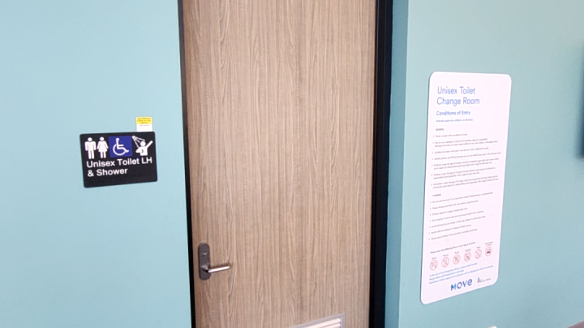 Wood-grain door with silver lever handle and vent. Black sign on blue wall says 'Unisex Toilet LH & Shower' with icons. Large white 'Conditions of Entry' sign is mounted on the wall to the right.