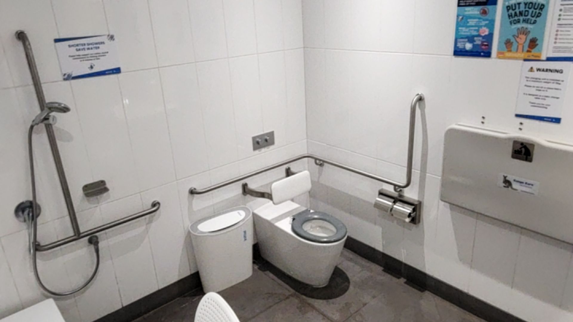 Accessible bathroom with white tiled walls and level grey floor. Includes a toilet with backrest, stainless steel grab bars, handheld shower on rail, and wall-mounted fold-down baby change table.